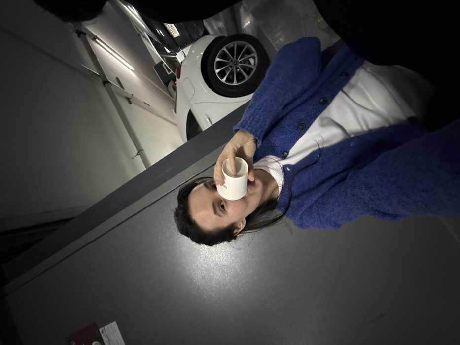 The image depicts an indoor parking garage in Tel-Aviv, Israel. A female subject with dark hair is visible in the foreground, facing the camera. She wears a blue, textured cardigan over a white shirt and is holding a white disposable cup with "HOLMES" printed on it to her mouth, appearing to drink. The top of a second individual's head, dressed in dark clothing, is partially visible on the far right. The immediate background features a dark gray wall with a red fire alarm call point and a diagonal blue-grey structural beam. Further back, two parked vehicles are visible: a white hatchback on the left and a dark sedan on the right. The dark sedan displays an Israeli license plate with the partial sequence "310-XX-XXX". Overhead, typical fluorescent garage lighting illuminates the scene. The presence of the "HOLMES" branded cup and the Israeli license plate suggests the location is a parking garage associated with a Holmes Place facility in Tel-Aviv.