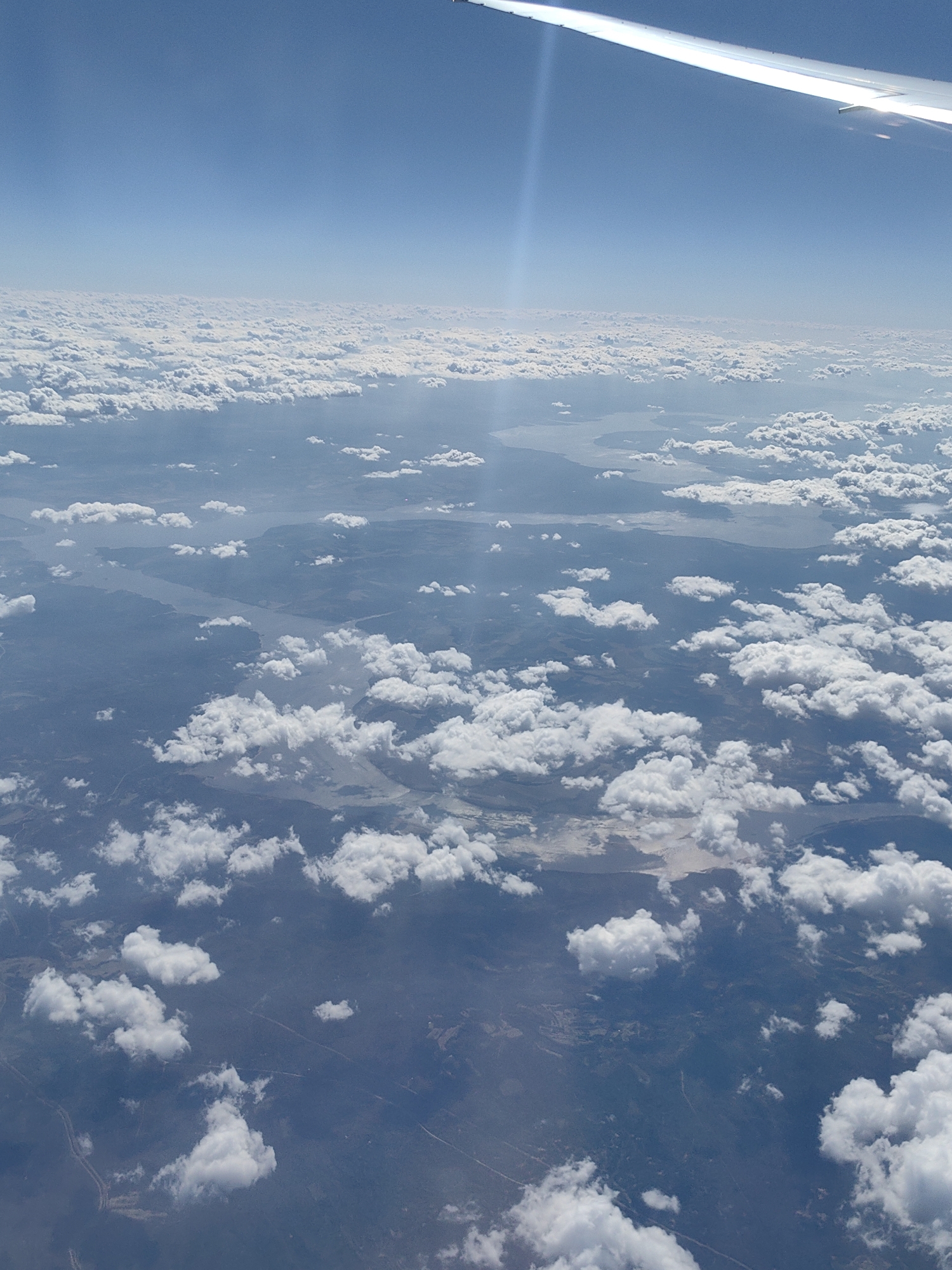 The image shows an aerial view from an airplane window. Above, there's a clear blue sky, transitioning to a blanket of clouds on the horizon. Below the clouds, the landscape is visible with patches of water bodies and land, partially obscured by scattered clouds. The wing of the airplane is seen at the top right corner. There is no data to suggest the location is Khimki, Russia.