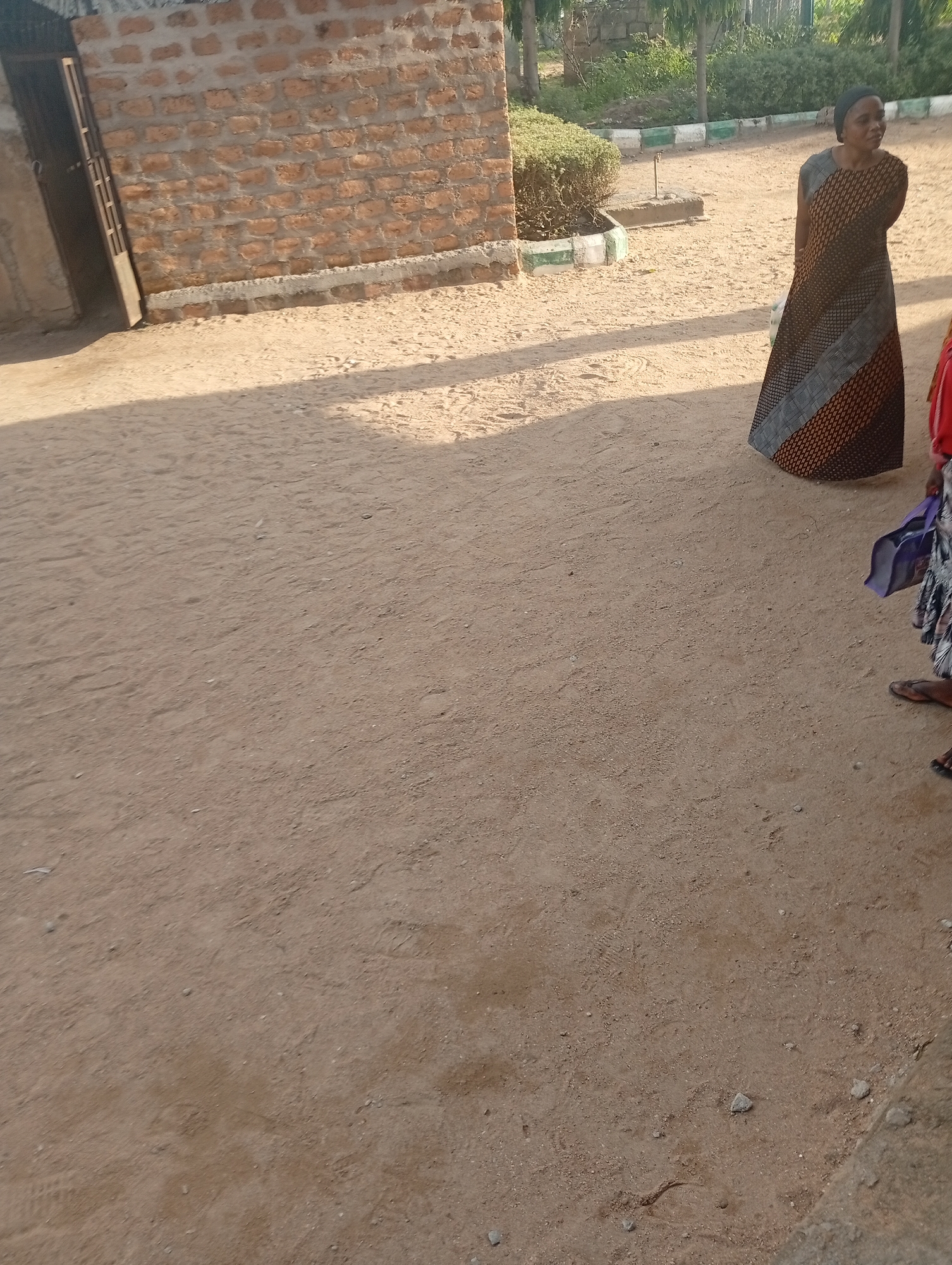 The image shows an outdoor scene in Jalingo, Nigeria. A brick building with a metal gate is visible on the left. In the center and right of the image, there are two women. One woman, on the right, is wearing a patterned dress and a headscarf, and appears to be looking to the side. Another woman, partially visible on the far right, is wearing a patterned skirt and holding a purple bag. The ground is covered in sand, and there are some low bushes and trees in the background, suggesting a sunny day. The lighting and shadows indicate it is likely late afternoon. The overall impression is of a casual, everyday scene in a Nigerian town. There is no visible text in the image.