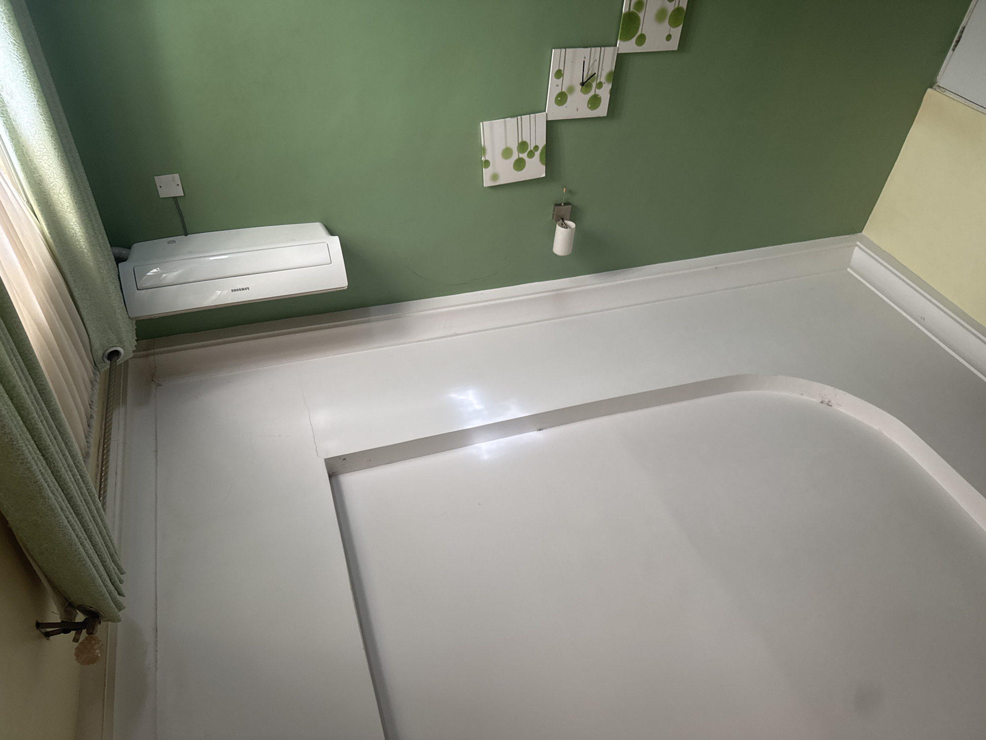 The image depicts an interior scene, likely a room in a house in Narayi, Nigeria. The dominant colors are a muted green on the wall and a bright white on a large, flat surface occupying the lower portion of the frame. Mounted on the green wall is a white air conditioning unit, brand name "SAMSUNG" visible. Above the air conditioner, a small white electrical outlet with a grey wire extending from it is visible. On the green wall to the right, a decorative clock composed of three square panels with a green dot pattern and clock hands is mounted. Below the clock, a small white toilet paper holder with a roll of toilet paper is attached to the wall. The transition between the green wall and the white surface is marked by a white trim. The white surface appears to be a raised platform or a large piece of furniture with a curved edge. To the far left, a light green curtain is partially visible, draped and held by a metal fixture. The lighting suggests it is daytime, with light filtering in from an unseen window, casting reflections on the white surface. There are no people visible in the image. The overall impression is of a clean, modern, and possibly residential space.