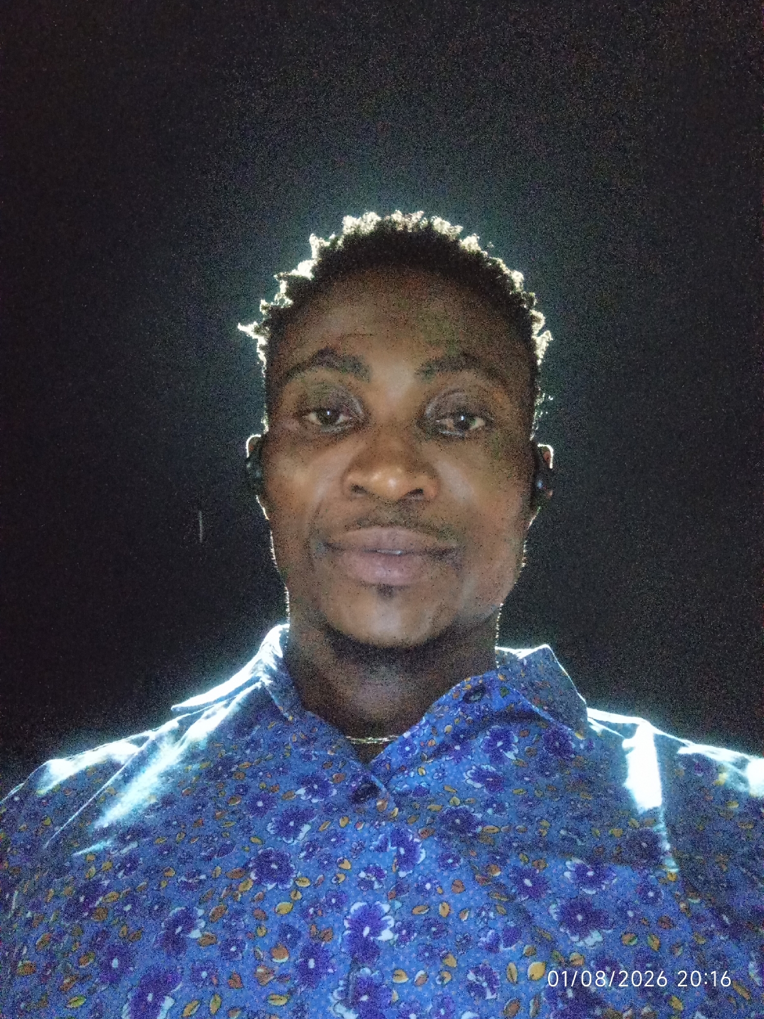 A medium shot, head-on portrait captures a Black male in Adankolo, Nigeria, against a dark, grainy background. The man has short, tightly curled hair that is highlighted by a strong light source from above, creating a halo effect. He has dark eyes, a broad nose, and a goatee. He is wearing a blue button-down shirt with a floral pattern of purple flowers and yellow and orange accents. A faint chain is visible around his neck. The lighting is dramatic, with a strong beam illuminating his face and shoulders from the front and slightly from the side, casting subtle shadows. The timestamp "01/08/2026 20:16" is visible in the bottom right corner of the image.
