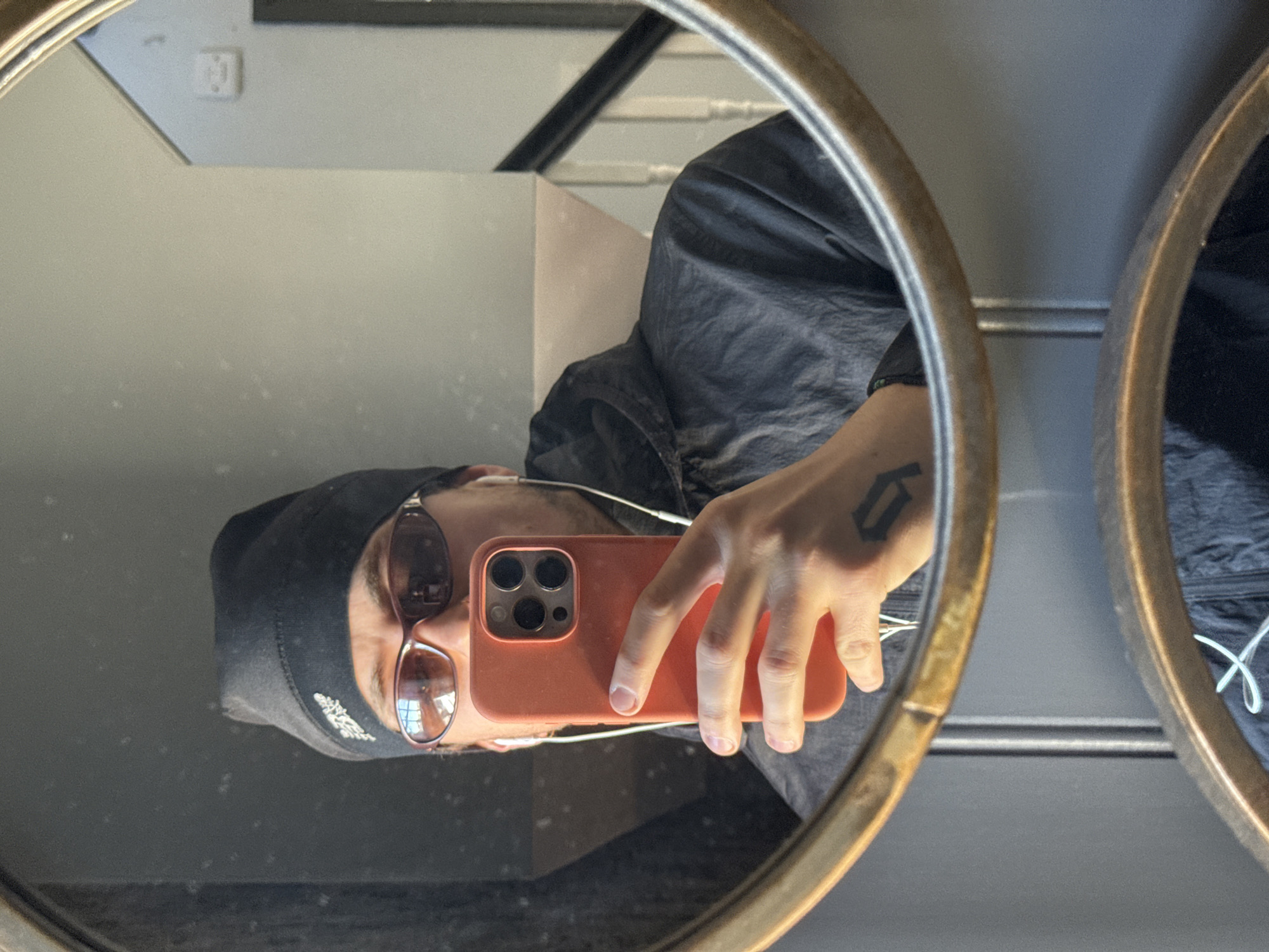 The image is a mirror selfie, rotated 90 degrees counter-clockwise, showing a person's reflection. The individual, appearing horizontal in the frame, is taking the photo with a smartphone.
The subject is wearing a dark cap with an indistinct white logo or lettering, brown-tinted sunglasses, and a dark, possibly grey or black, jacket. White wired earphones are visible, leading from their ears towards the smartphone. Their left hand, which is holding the phone, has a tattoo on the back, depicting a stylized block letter that resembles a 'G' or a '6'. The smartphone is an iPhone model with three cameras, enclosed in a bright orange protective case.
The selfie is captured in a round or oval mirror with a distinctive golden or brass-colored frame. The reflection offers a glimpse of the indoor setting: a light-colored wall behind the subject, featuring a light switch or electrical outlet in the upper-left of the reflection. A dark, possibly black, railing or beam is also visible, suggesting a modern interior. Part of another similar mirror or reflective surface can be seen on the far right.
The scene appears to be well-lit, likely during the daytime, with bright reflections visible in the sunglasses, possibly from windows or overhead lights. The general environment suggests an indoor location, consistent with residential or commercial architecture. The activity is clearly a self-portrait taken via a mirror. The location context is Stotfold, United Kingdom.