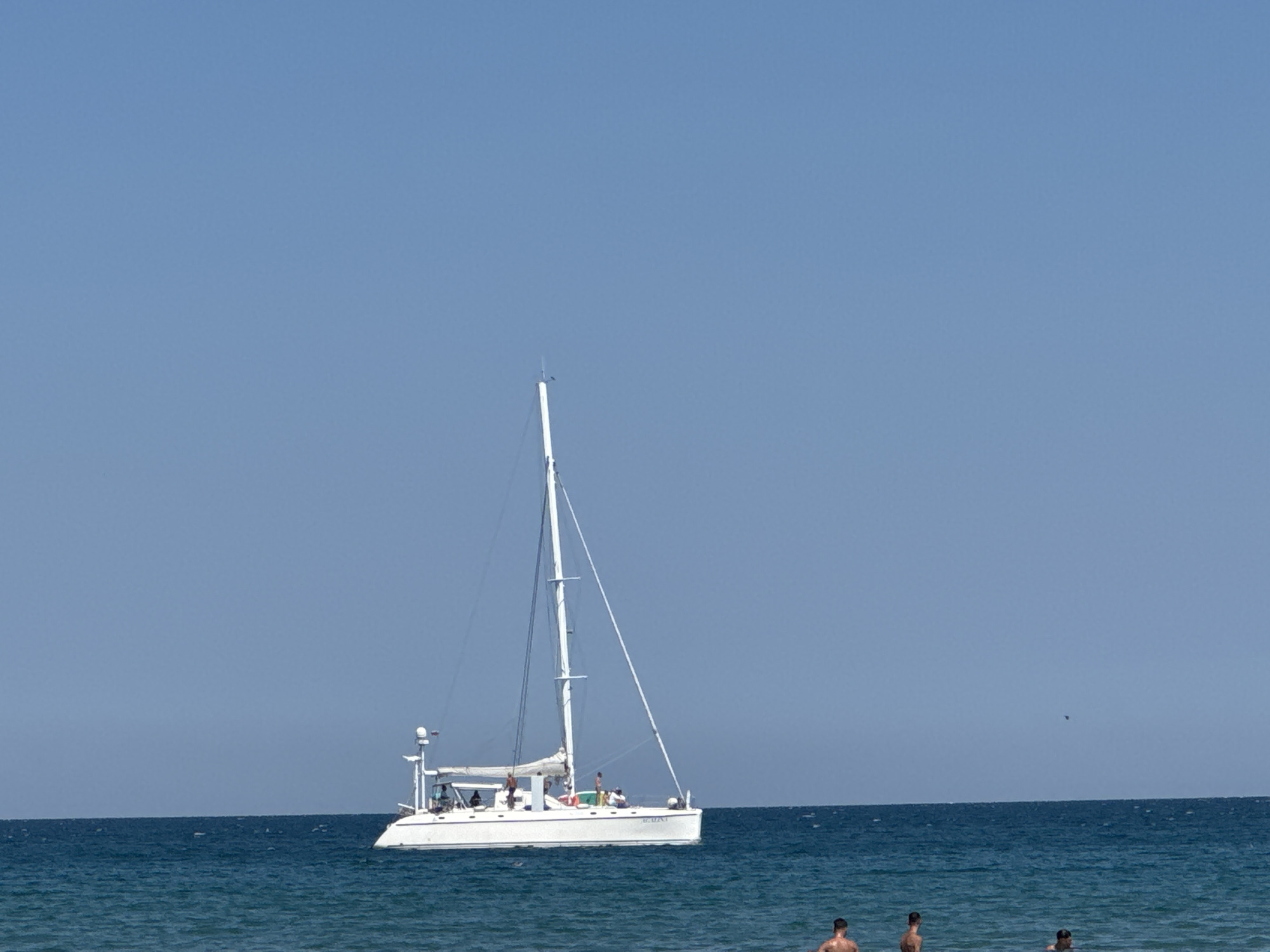 In the city of Sozopol, Bulgaria, the image depicts a bright, clear day at the beach. The dominant element is a white catamaran sailing on the deep blue sea. The catamaran has two masts and several people are visible on its deck. The horizon line is clearly defined between the sea and the vast, light blue sky, which occupies the majority of the frame. In the foreground, partially submerged in the shallow waters, are three men, their torsos visible. The overall scene conveys a sense of leisure and a typical summer day by the Black Sea coast.