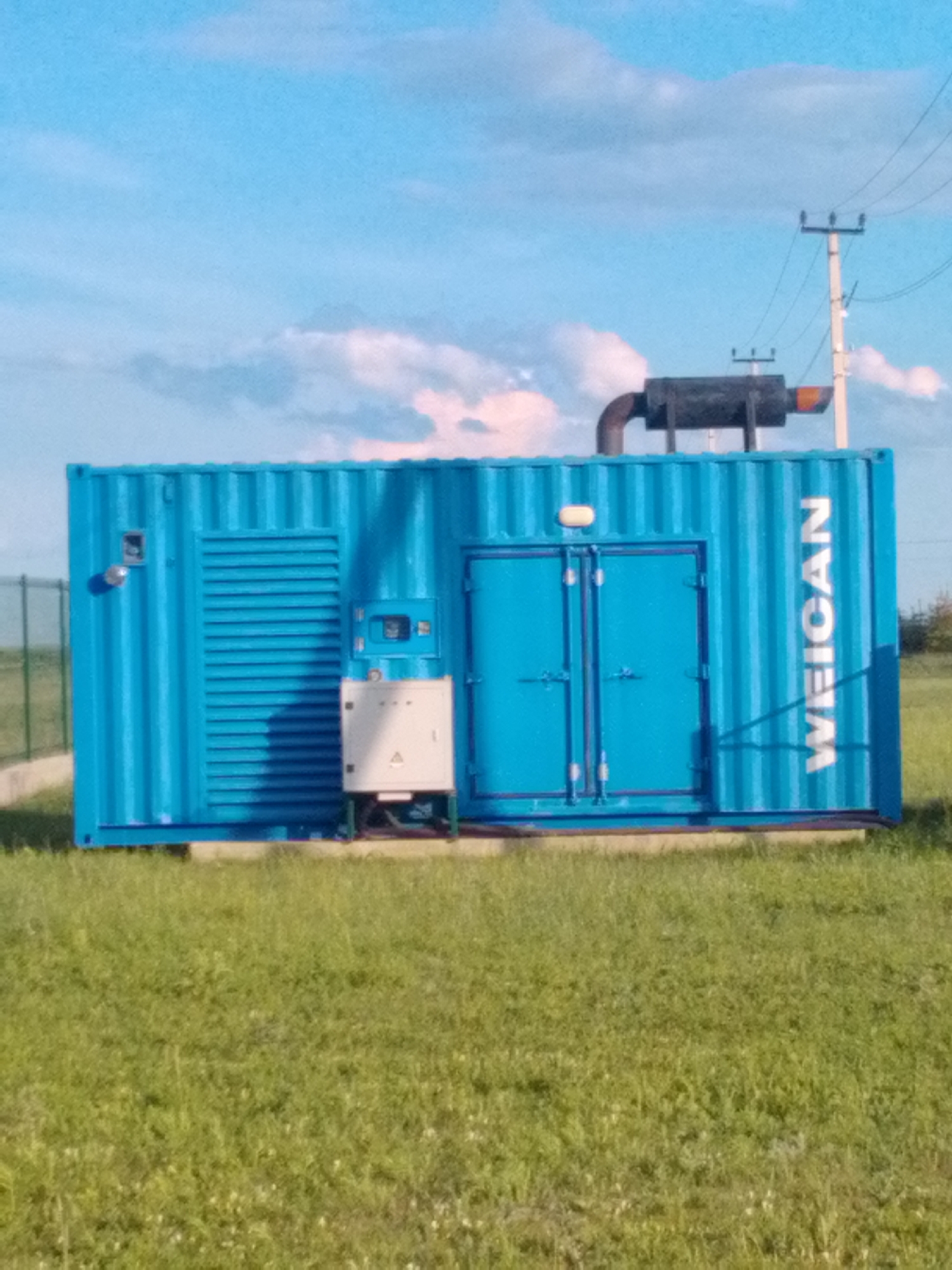 The media file shows a blue shipping container unit, likely a generator or power supply, placed outdoors on a grassy field. The container is rectangular with ribbed sides and two large doors on the front. A smaller white electrical box is mounted on the left side of the container. On top of the container, a black exhaust pipe with a curved section is visible. The background features a bright blue sky with scattered white clouds. A utility pole with wires is visible in the upper right corner. The setting appears to be a rural or semi-rural area, given the open field and the presence of basic infrastructure like the utility pole. The lighting suggests it is daytime, likely late afternoon or early morning due to the soft light and shadows. The image is a still shot, and no activity or people are depicted. Visible text on the right side of the container reads "WEICAN" in large white letters. The context provided places the scene in "Корнеев ауылдық округі, Kazakhstan."