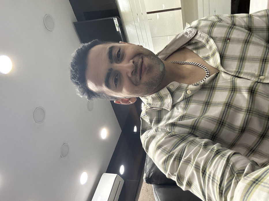 A man with dark hair and a slight smile wears a plaid shirt and a silver chain. He's positioned at an angle, facing the camera. The setting appears to be indoors, with a ceiling featuring multiple recessed lights. A dark-colored TV screen and some furniture are visible in the background.