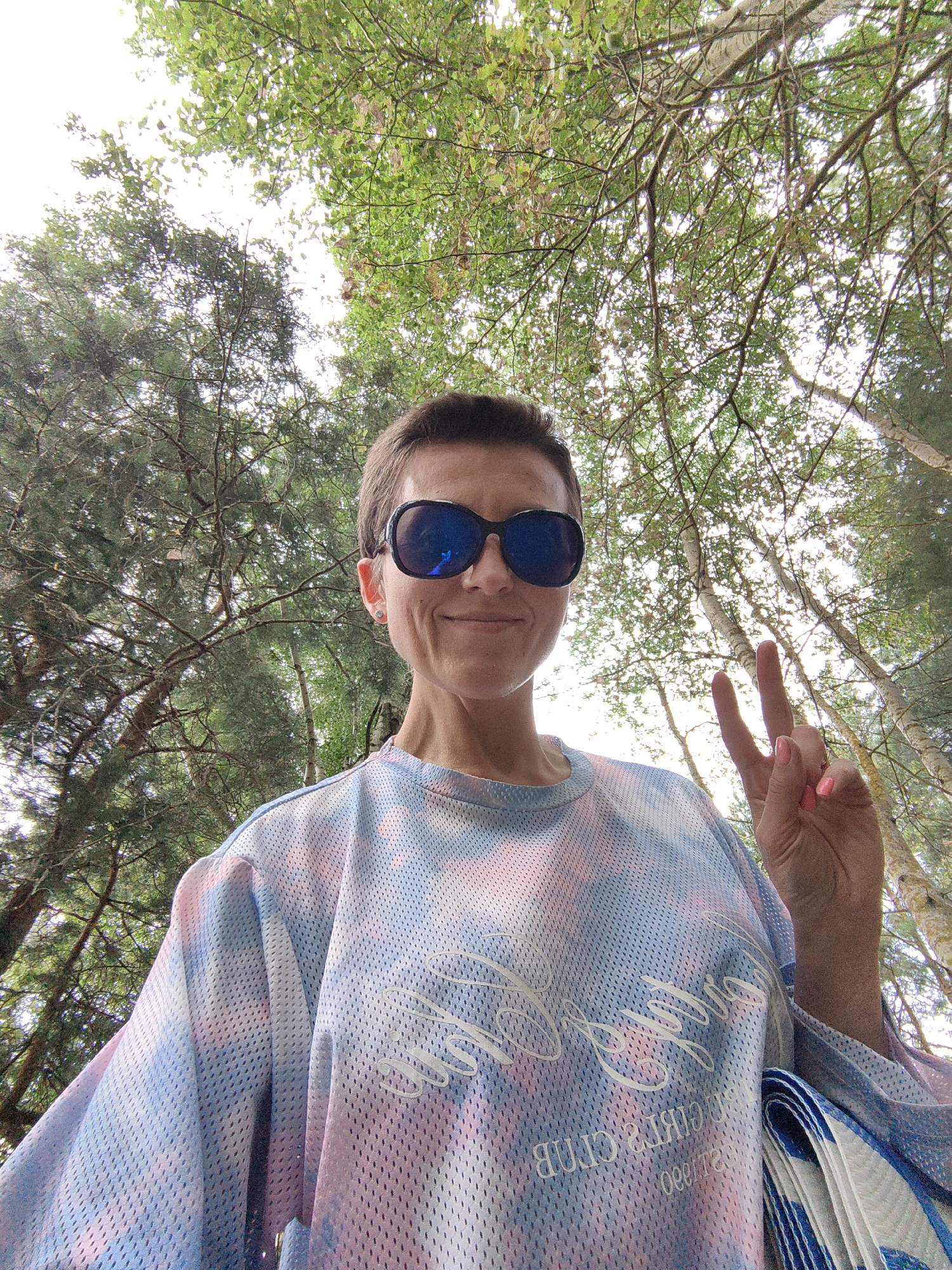 A woman with short brown hair and sunglasses is taking a selfie outdoors. She is wearing a tie-dye t-shirt with blue, pink, and white colors. Her right hand is raised, and she is making a peace sign with her fingers. The background consists of trees with green leaves and a bright sky. The location is identified as Myadzyelski Sil'ski Savet, Belarus.