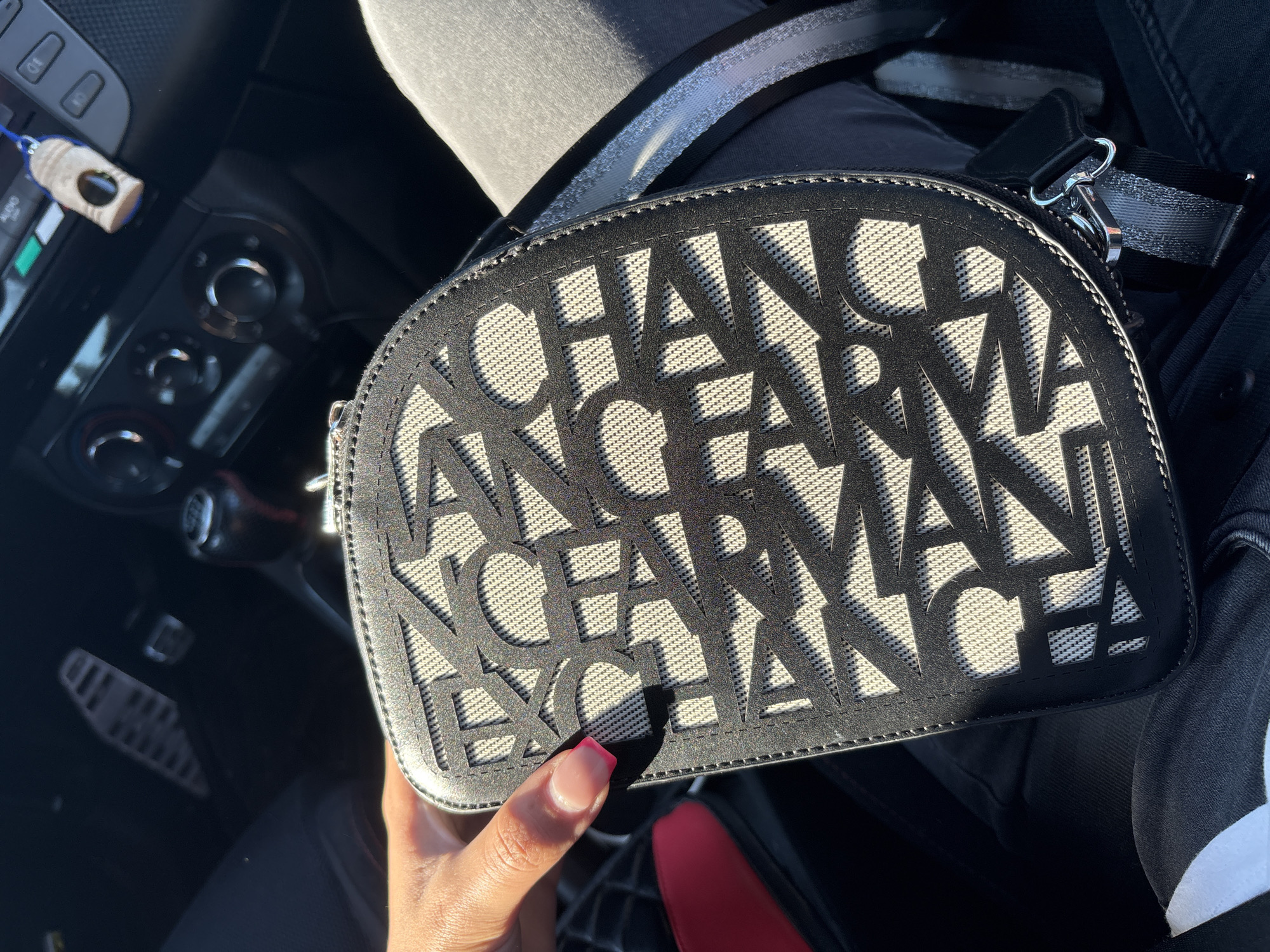The image shows a woman's hand holding a black and white patterned bag. The bag has a geometric design with cutouts that reveal a textured fabric underneath. The hand has neatly manicured nails with a pink and white French manicure. The background appears to be the interior of a car, with a gear shift and dashboard visible. The scene is outdoors as indicated by the sunlight casting shadows on the bag. The location of Vratsa, Bulgaria is not evident from the image.