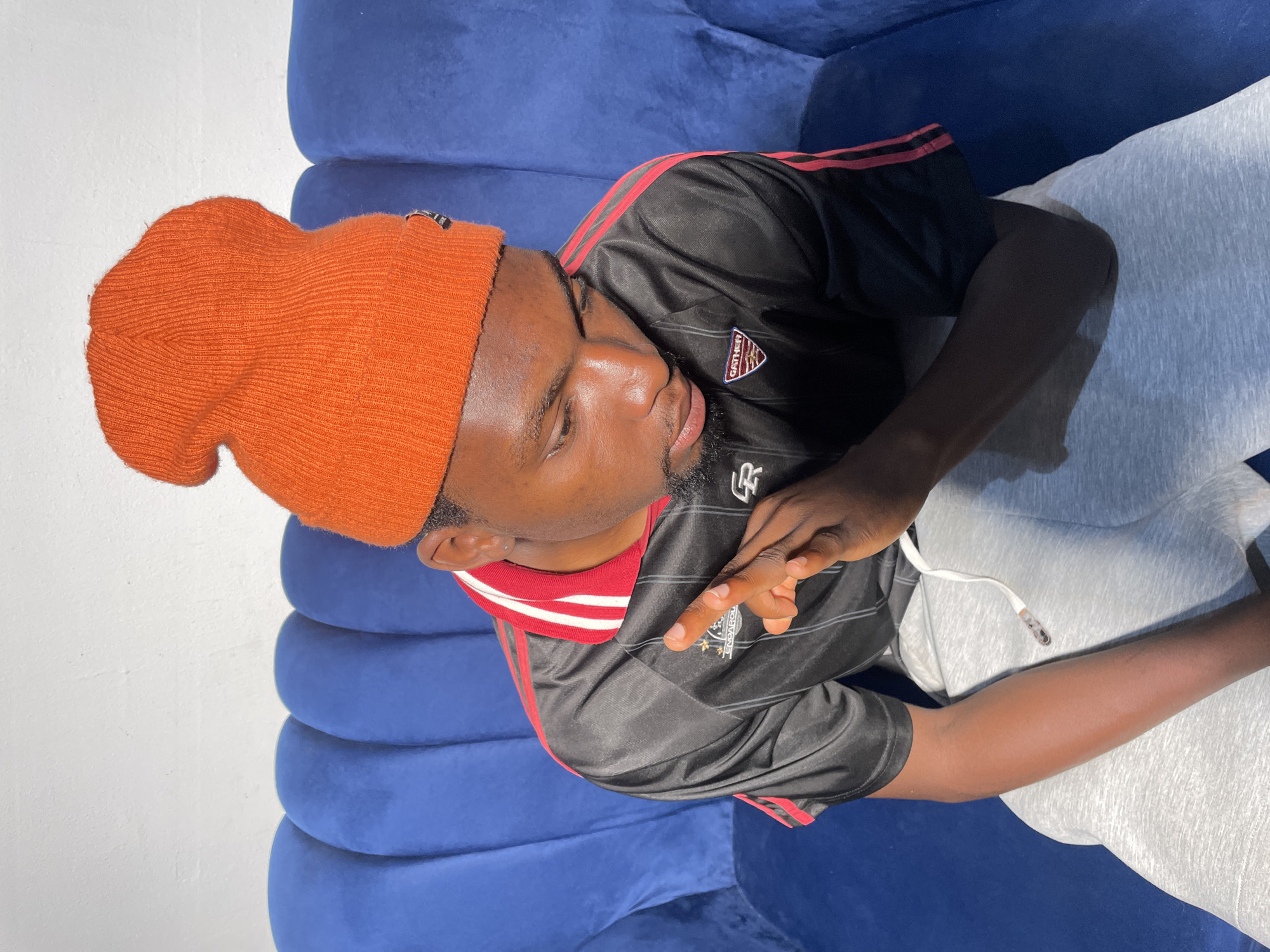 A young Black man is seated on a blue couch. He is wearing an orange beanie, a black shirt with red stripes on the sleeves, and gray sweatpants. He is making a hand gesture. The location is not identifiable as Barakin Akawo, Nigeria from the given image.