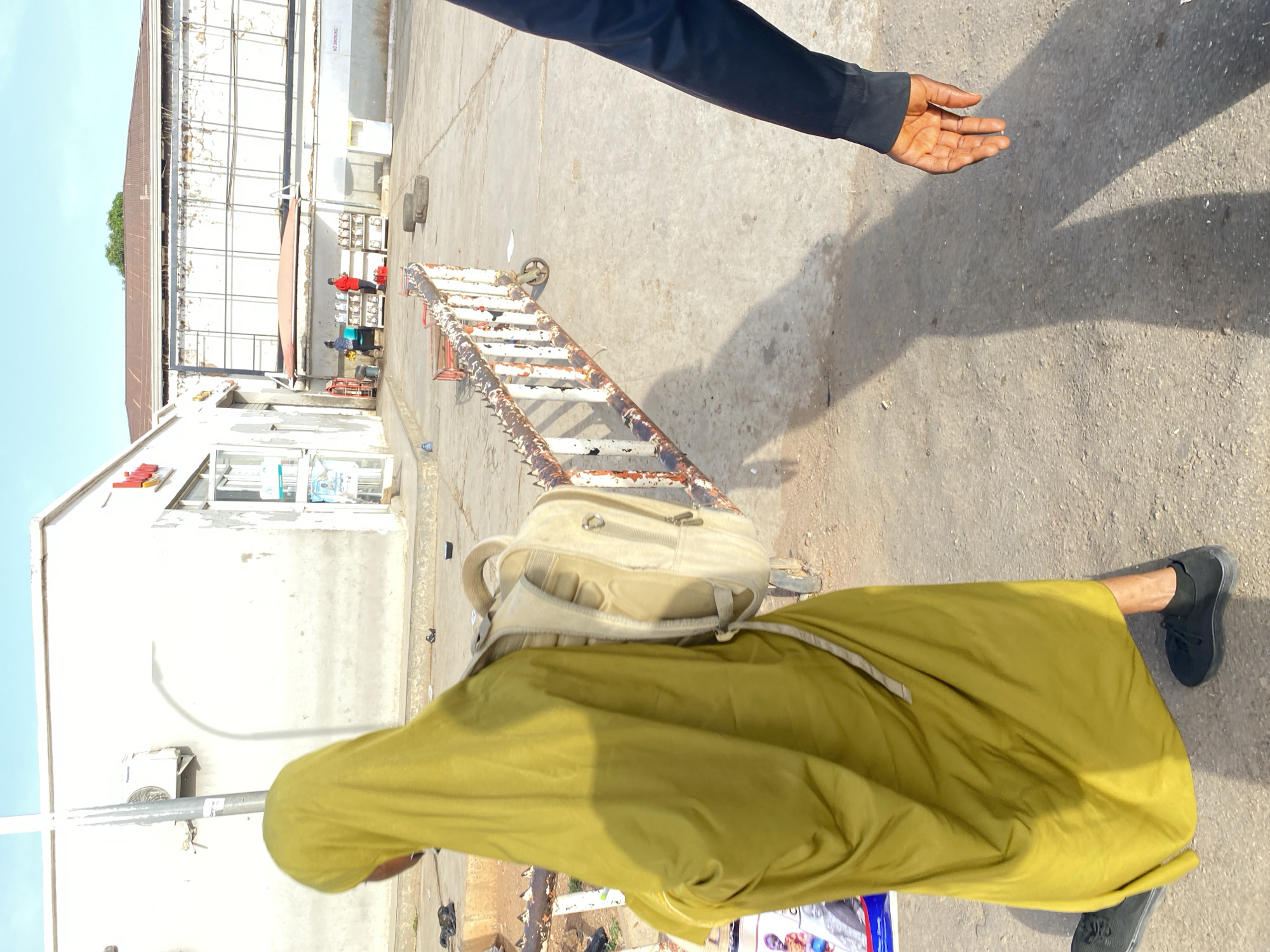 A medium shot, captured at eye-level, shows a person in a yellow robe and backpack walking on a paved road in Jos, Nigeria. The person is wearing black shoes. Another person in a dark-colored jacket with his hand partially visible is walking by. A metal ladder is lying horizontally on the road. In the background, there is a white, one-story building with red signage, a metal structure behind it, and two people by a table. The sky is clear. The lighting suggests a sunny day.