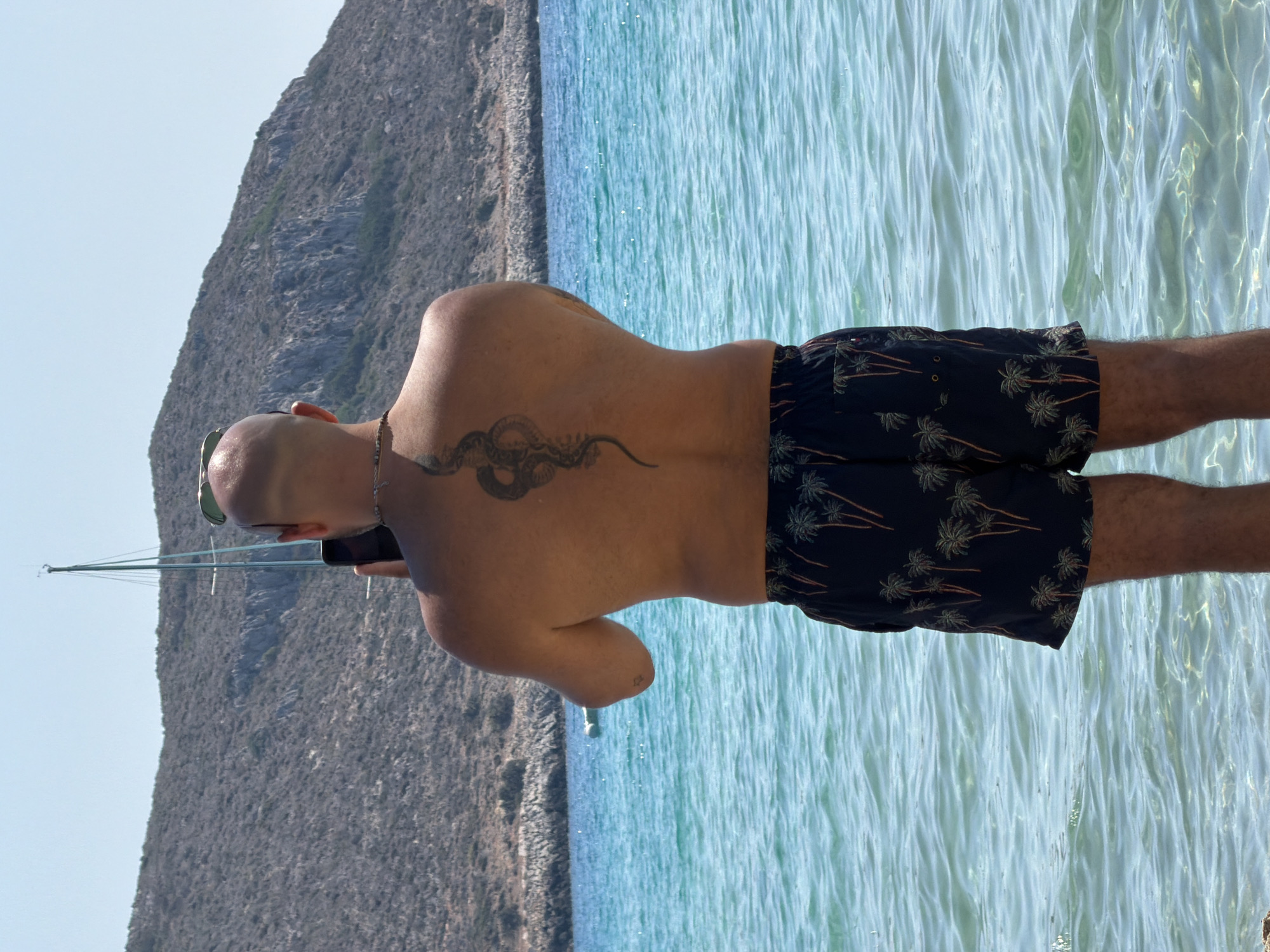 A man with a bald head and a snake tattoo on his back stands in the shallow water of a turquoise sea, facing away from the camera and holding a smartphone. He wears dark swimming shorts patterned with palm trees. In the background, a sailboat with a tall mast is visible on the water, and a rocky, arid hillside rises behind it under a clear blue sky. The water is calm with gentle ripples.