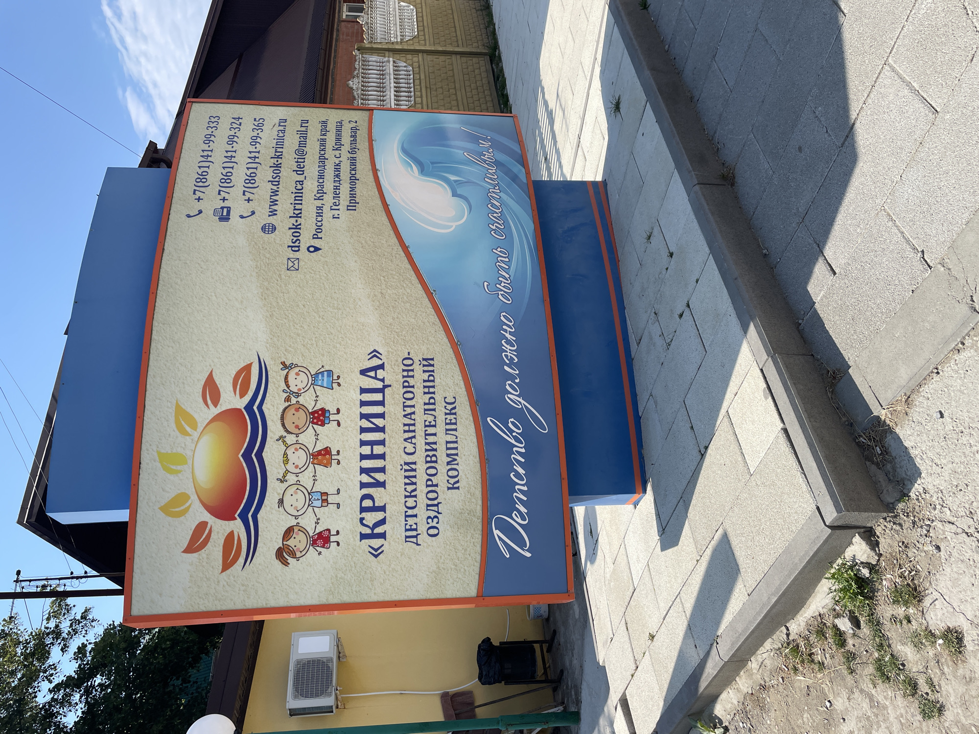 A large, colorful billboard advertising "KRINitsa" Children's Sanatorium and Health Complex stands on a paved sidewalk in Krinitsa, Russia. The billboard features cartoon drawings of children and a sun rising over waves. Contact information, including phone numbers, website, email address, and physical address, is displayed on the right side. The physical address provided is Primorsky Boulevard, 2, Gelendzhik, Krinitsa, Krasnodar Krai, Russia. The billboard is supported by a blue and orange structure. In the background, a yellow building with an air conditioning unit is visible, along with a black trash can and some greenery. The sky is clear and blue, indicating a sunny day.