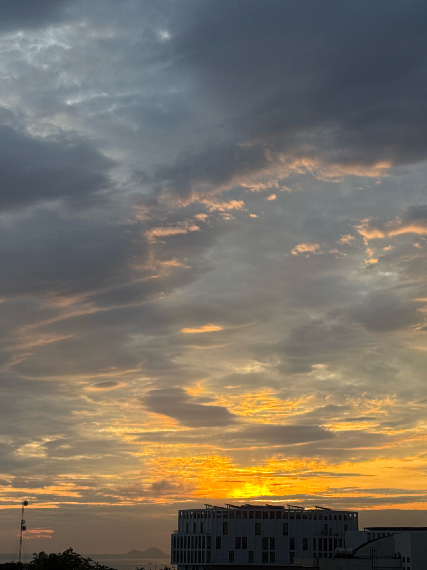 A wide-angle shot captures a dramatic sunset over Nha Trang, Vietnam. The sky is filled with layers of clouds, ranging from dark gray to vibrant orange and yellow hues, illuminated by the setting sun. In the foreground, a modern white building with a distinctive lattice-like rooftop is visible. To the left, a tall antenna-like structure with multiple platforms stands against the horizon. The distant ocean is calm, with a faint outline of islands visible in the far distance. The overall scene evokes a sense of twilight tranquility.