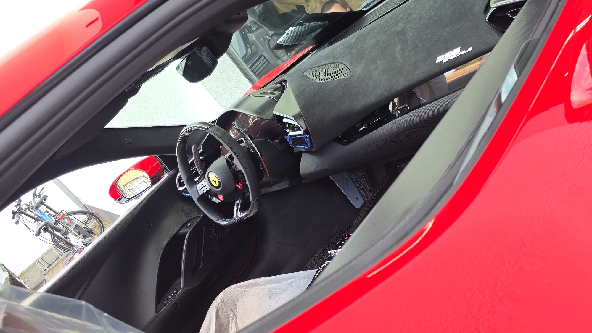 The image provides an interior view of a red sports car, looking towards the driver's side. The car's door is open, revealing the steering wheel, dashboard, and part of the seats. The steering wheel features the Ferrari logo. The interior appears to be made of black materials, possibly leather and carbon fiber. Outside the car, through the open door and windshield, a bicycle is visible mounted on a rack, suggesting the car might be parked in an urban environment or near a residential area. The lighting suggests it's daytime.