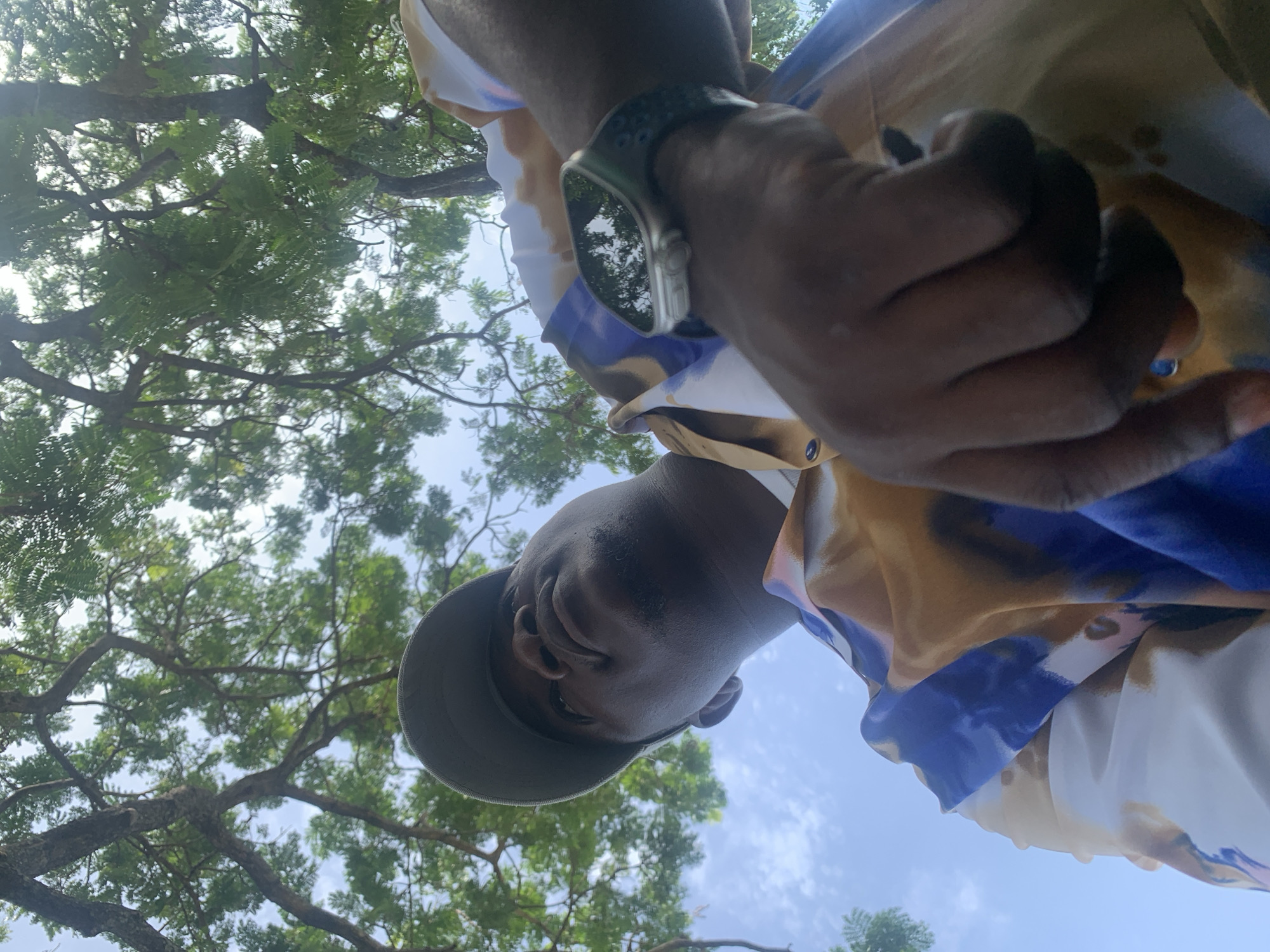 A low-angle shot looking up at a Black man wearing a light grey cap, a smartwatch on his left wrist, and a patterned shirt featuring blue, white, and yellow hues. He appears to be holding something in his hand, but it is out of focus. The background consists of a tree with dense green foliage, a clear blue sky visible through the branches. Given the prompt, the location would be Akwanga, Nigeria.