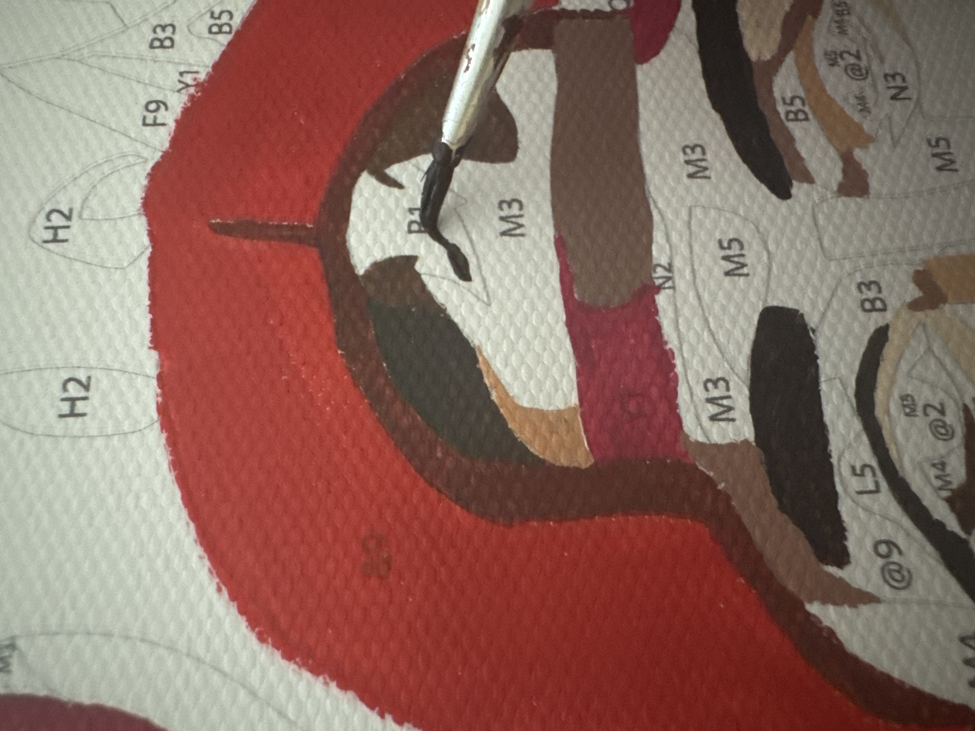 This close-up image captures a segment of a paint-by-numbers project in progress in Chaikovsky, Russia. A paintbrush, featuring a silver ferrule and black bristles, is actively applying a dark color to an unpainted section of the textured canvas. The canvas surface is visible with its distinct weave.
Portions of the artwork are already filled with various shades of paint, including a dominant dark red, dark brown, medium brown, light tan, and a deep reddish-brown. Unpainted areas display intricate outlines, within which alphanumeric and special character codes are printed, guiding the painter on which color to apply next.
Notable visible text and codes on the canvas include: H2, F9, Y1, B3, B5, P1, M3, N2, M5, L5, @9, M4, @2, MB5, and N3. The specific subject of the painting is not entirely clear from this cropped view, but the different colored shapes suggest a detailed image is taking form. The lighting indicates an indoor setting.