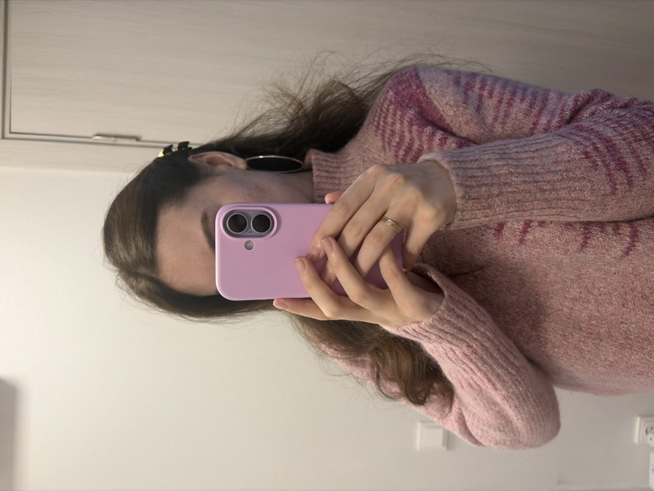 A person holds a light purple phone, possibly taking a selfie. They wear a pink sweater with a textured pattern. The individual has brown hair, styled with a clip and hoop earrings. A simple gold ring adorns their finger. The background features a white wall with a cabinet door visible in the upper left and an electrical outlet in the lower right. The image appears to be taken in a bathroom or similar indoor setting.