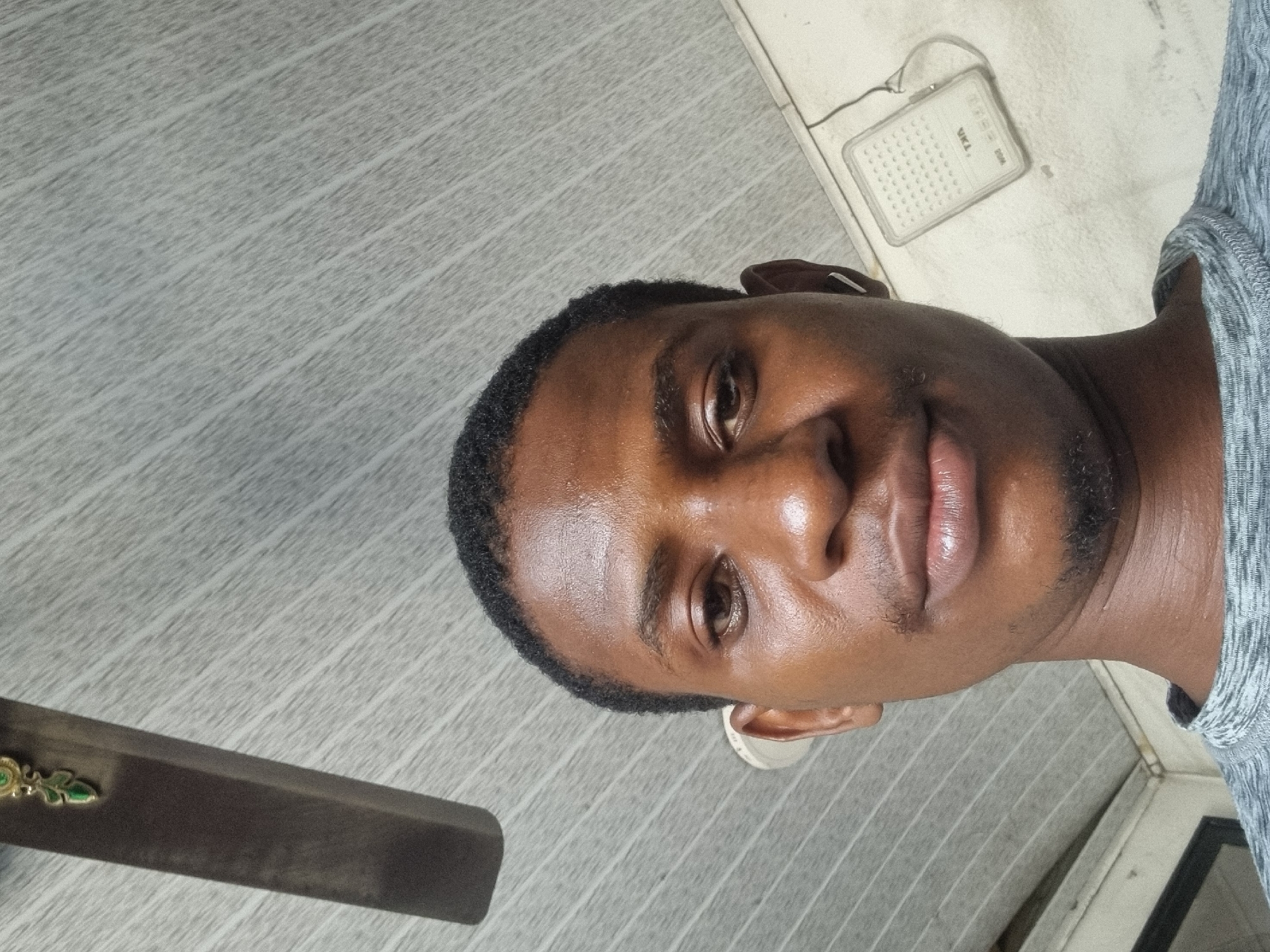 The media file presents a close-up portrait of a young man, likely taken indoors in Onitsha, Nigeria. The subject, a man with dark skin, short dark curly hair, and light stubble on his chin and jawline, looks directly at the camera with a neutral to slightly serene expression. His skin has a visible sheen, indicating good lighting. He is wearing a textured, light grey long-sleeved shirt.
The background is a light grey, textured surface with diagonal linear patterns, resembling a tiled or paneled wall or ceiling. In the upper right portion of the frame, behind the man's head, there is a small, white rectangular electronic device. This device, possibly a portable radio or speaker, features a grid of small holes and discernible text that reads "VKA" and "2004". A thin wire is also visible extending from it. In the lower left corner of the image, a dark, metallic object with a small, ornate green and gold emblem is partially visible. The lighting is soft, emphasizing the man's features. No specific activity is depicted beyond posing for the photograph.