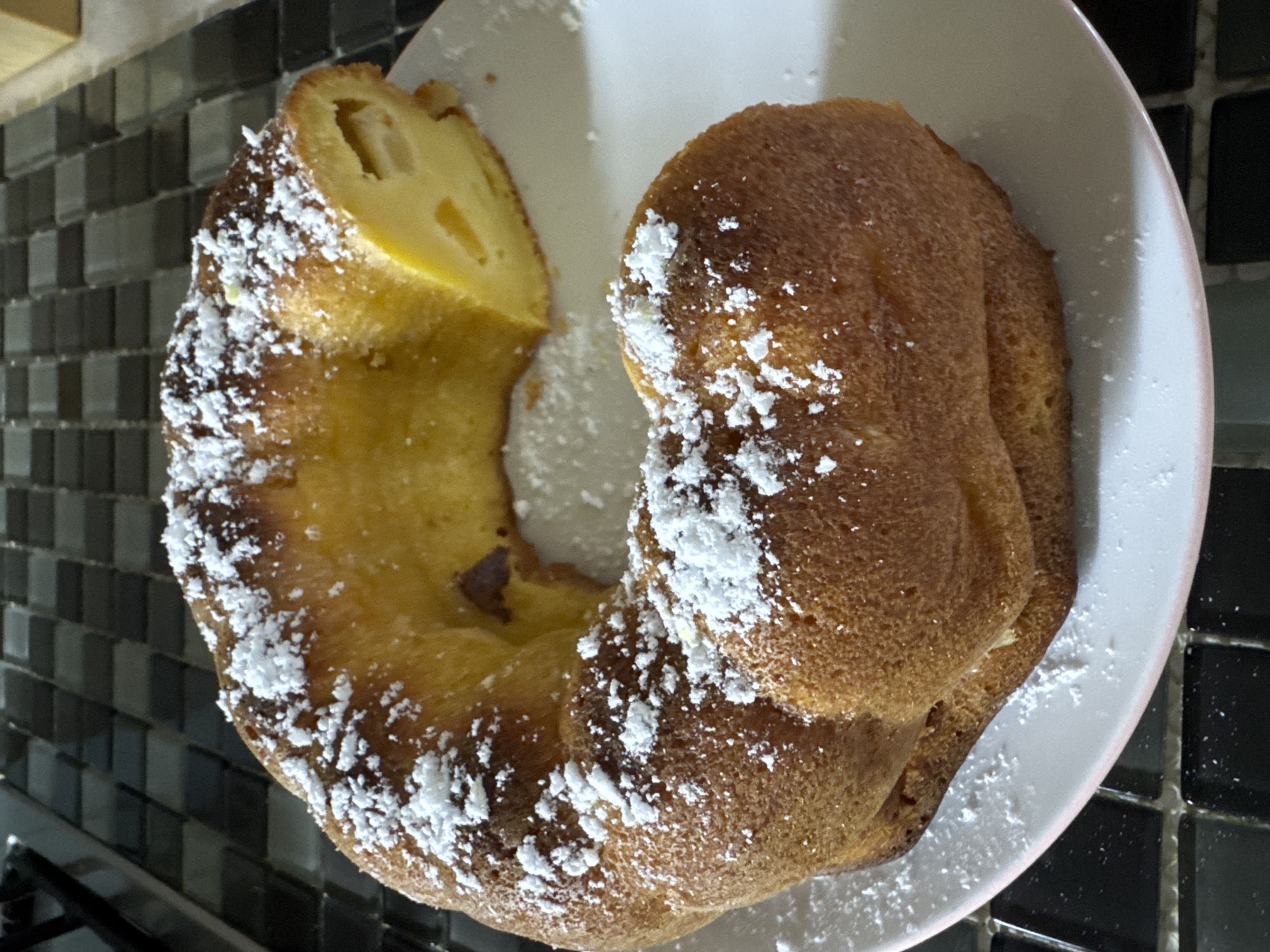 A bundt cake dusted with powdered sugar sits on a white plate. A slice has been removed from the cake, revealing a light yellow interior with chunks of fruit. The plate is on a black and white tiled countertop. The cake is the primary subject. The scene is domestic and appears to be a kitchen.