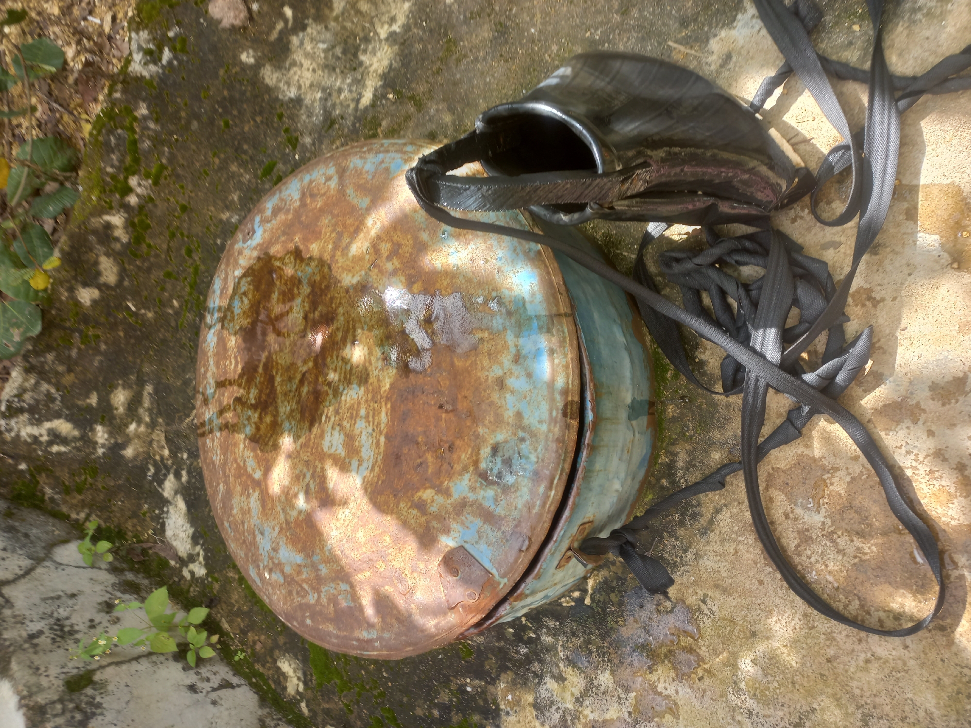 In Jos, Nigeria, a rusty, circular, blue and brown metal drum with a lid sits on a weathered concrete surface. A black, braided strap, possibly from a bag, is draped around the drum and lies coiled on the ground. A black bag, made of a shiny, possibly rubber material, is also present, positioned next to the drum. The surrounding environment shows signs of moss and some greenery, suggesting an outdoor setting that may be somewhat neglected. The concrete is damp in places, indicating recent rainfall or moisture.