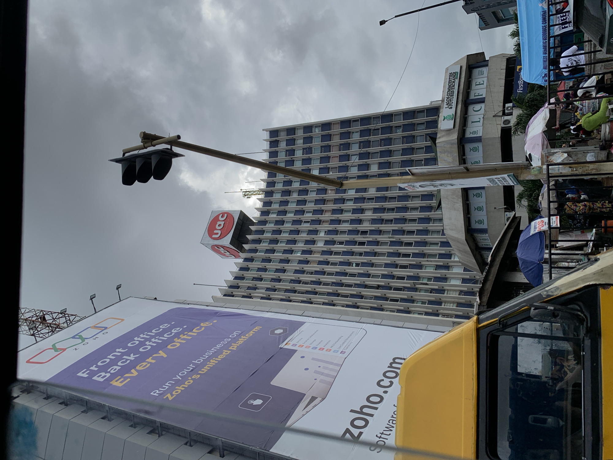 The image captures a vibrant street scene in Lagos, Nigeria, under an overcast sky. In the foreground, partially visible on the bottom right, is the yellow cab of a vehicle, possibly a bus or truck, indicating a street-level perspective. A prominent element on the left is a large billboard advertising "ZOHO" software, with text reading "Front office. Back office. Every office. Run your business on Zoho's unified platform," along with "zoho.com" and various software categories like "Service," "Email & Collaboration," and "Analytics."
Dominating the mid-ground is a tall, multi-story building featuring a distinctive blue and white checkerboard facade of windows. Near its top, a red "uac" logo is visible. To the right of this tall building, another building houses the "Lagos Commodities & Futures Exchange" (LCFE), with its name clearly displayed on its exterior. Further to the right, a "FirstBank" sign is also partially visible.
The street level to the right of the buildings appears bustling with activity. People are gathered among what seem to be market stalls, some shielded by colorful umbrellas, including a blue one, suggesting a commercial or market area. Banners with text like "EXPRESS POS" are also present. A traffic light fixture is suspended across the top-center of the image, indicating an intersection or busy road. The overall impression is that of an active urban environment.