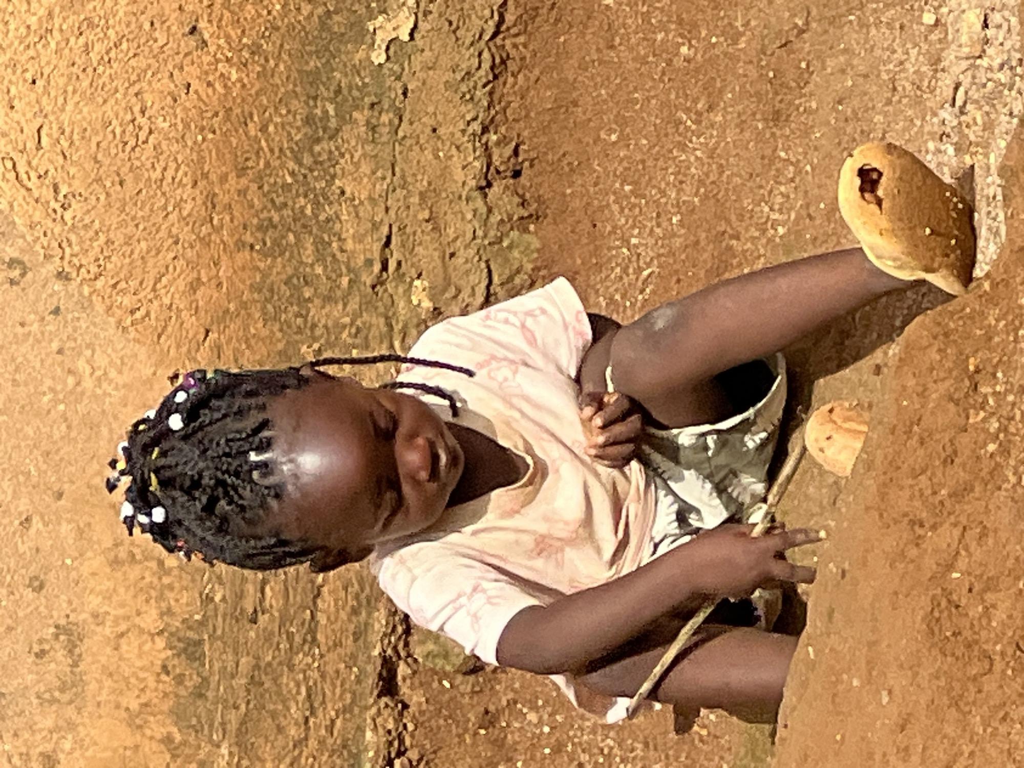 A young Black girl with braided hair adorned with colorful beads is sitting in dirt. She is wearing a light-colored t-shirt with a pattern and patterned shorts. She is holding a stick and appears to be digging or playing in the dirt. Her left foot is resting on a flat, wooden sandal. The background consists of a rough, textured wall and sandy ground. The lighting suggests it is daytime. The scene is located in the city of Jos, Nigeria.