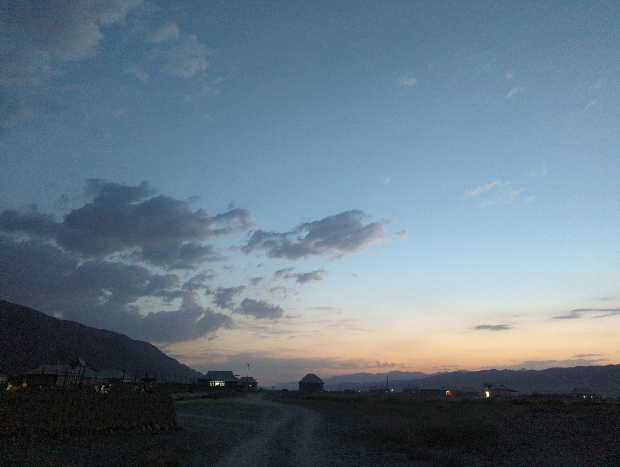 This is a wide-angle, eye-level, outdoor photograph taken during twilight in the village of Uch-Dobo, Kyrgyzstan. The sky is a gradient of pale blue to soft orange from top to bottom, with scattered dark and light gray clouds. In the foreground, a dirt road curves through a dark, uncultivated field. To the left, a large, dark mountain range dominates the landscape. Several small, dark-colored structures, some with lit windows, are visible in the mid-ground, scattered across the plain and nestled against the base of the mountain. Some figures, appearing as silhouettes, are standing outside one of the structures. The overall atmosphere is serene and rural, capturing the transition from day to night in a mountainous region.