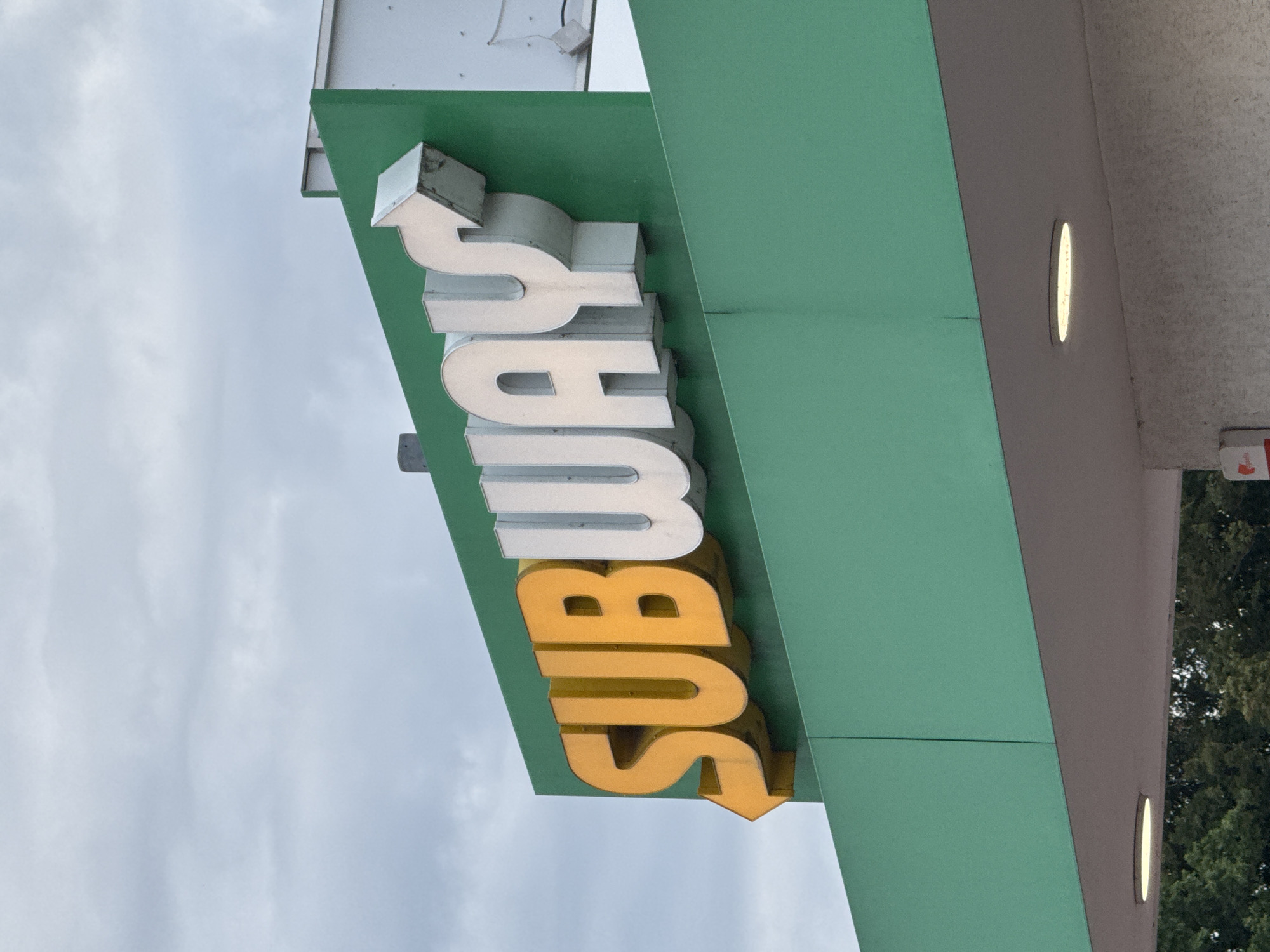A close-up shot captures a large, illuminated "SUBWAY" sign mounted on a green facade. The letters, some yellow and some white, are three-dimensional and cast shadows. An arrow, also three-dimensional, points left from the "S" in "SUBWAY". The sign is attached to a building structure with a white and grey exterior. The background consists of a cloudy, overcast sky. The scene appears to be outdoors, likely in an urban or suburban setting, given the building architecture.