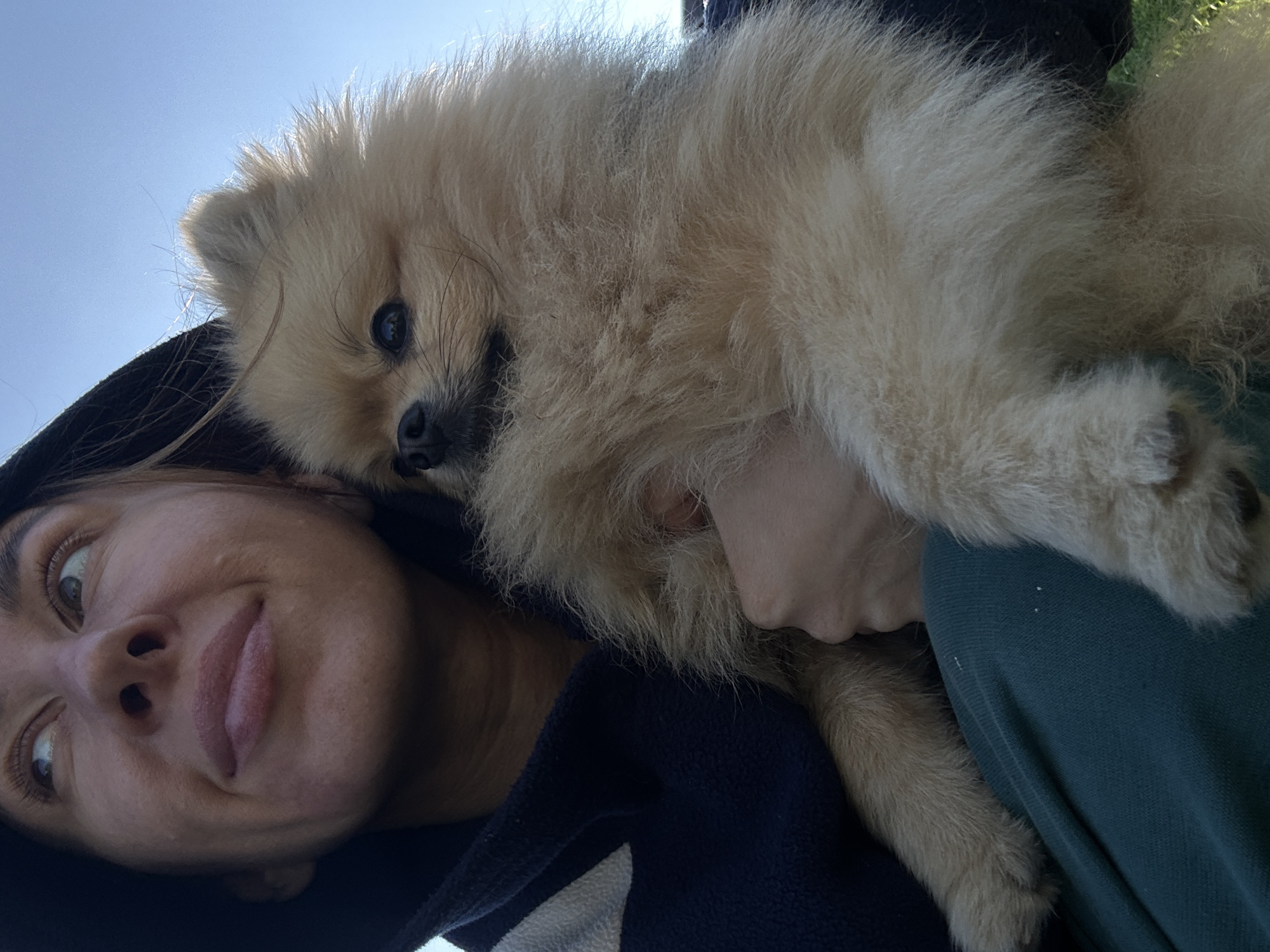 A woman, wearing a dark hooded jacket with a white stripe and dark green pants, holds a small, fluffy, light brown Pomeranian dog. The woman is looking upwards and to the left, with her mouth slightly open. The dog is nestled in her arms, facing towards the right side of the frame, with its eyes looking forward. The background consists of a clear blue sky. The scene appears to be outdoors, possibly in a park or open area, with a hint of green grass visible at the bottom right edge of the image. The specific location of Галево, Russia, is not depicted in the visual information provided.