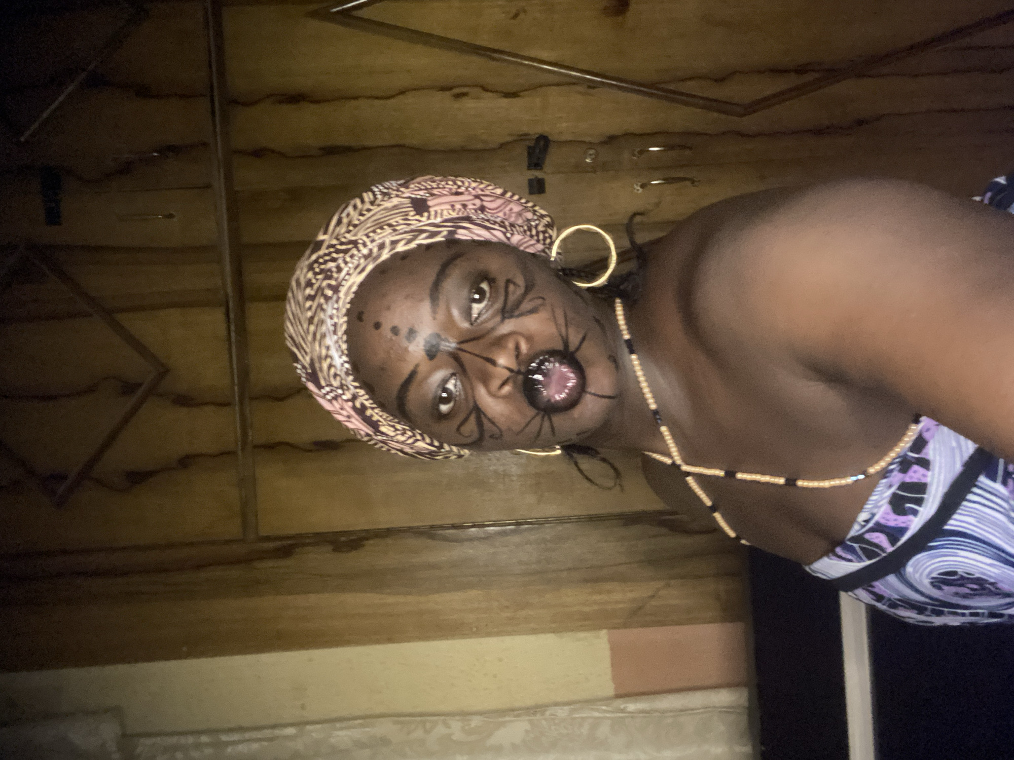 This media file captures a close-up portrait of an individual, likely a woman, with dark skin, positioned sideways in the frame. The subject is wearing an elaborate headwrap with intricate patterns in pink, brown, and cream tones. She is adorned with striking face paint, featuring dark, spider-like designs radiating from around her mouth and eyes, and several small dots on her forehead and cheeks. Her mouth is open, revealing her teeth and tongue, which also appears to have a dark design. She wears a large, gold-colored hoop earring in her right ear and a multi-strand beaded necklace with alternating light and dark beads that cross over her chest. Her attire includes a patterned top with blue, purple, white, and black hues.
The background suggests an indoor setting, possibly within a home or traditional structure in Maigizo, Nigeria. Wooden panels, possibly part of a wall or ceiling, are visible behind her, with some linear structural elements. The lighting appears dim, casting some shadows and highlighting the subject's face and jewelry. The overall impression is one of cultural expression or artistic performance, accentuated by the distinctive face paint and attire. There is no visible text in the image.
