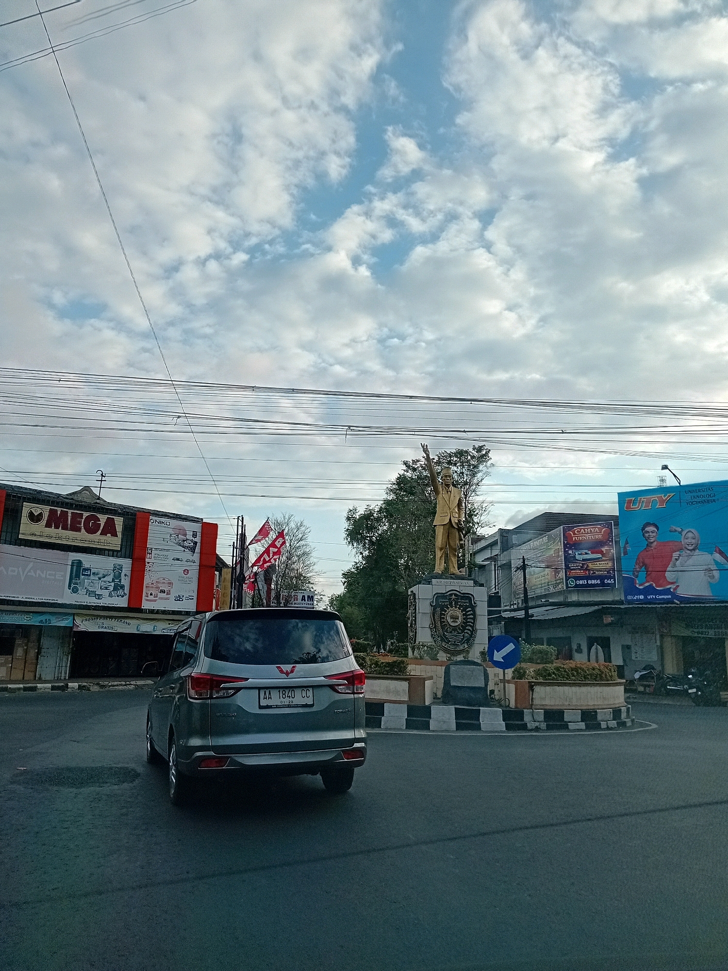 The image depicts a street scene in Purworejo, Indonesia, likely a city center or busy intersection. A silver minivan is positioned on the right side of the road, facing away from the viewer. In the background, a statue of a man in a golden suit stands on a pedestal in the center of a traffic circle. The statue's right arm is raised with the index finger pointing upwards. Surrounding the traffic circle are several buildings with signage, including "MEGA" and "UTY." A blue billboard featuring two individuals and the text "UTY" is prominently displayed. Red and white flags are visible on poles in front of some buildings. The sky is partly cloudy, with a mix of blue sky and white clouds. Power lines crisscross the upper portion of the image. The road surface is asphalt, and there are some plants and decorative elements around the traffic circle. The overall impression is of a lively urban environment.