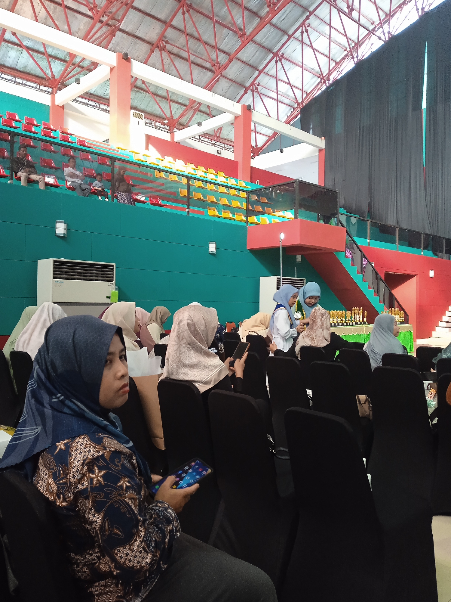 The image shows a gathering of women wearing hijabs inside a large, indoor venue in Pangongangan, Indonesia. The space features a raised seating area with red and yellow seats, a green wall, and a stage area draped with black curtains. Stairs leading up to the stage are visible. Numerous trophies are displayed on a table near the stage. The women are seated in black chairs, and one woman in the foreground is holding a smartphone. Some women are talking while others appear to be listening or watching. An air conditioning unit is mounted on the green wall. The structure of the building has visible red and white metal beams.