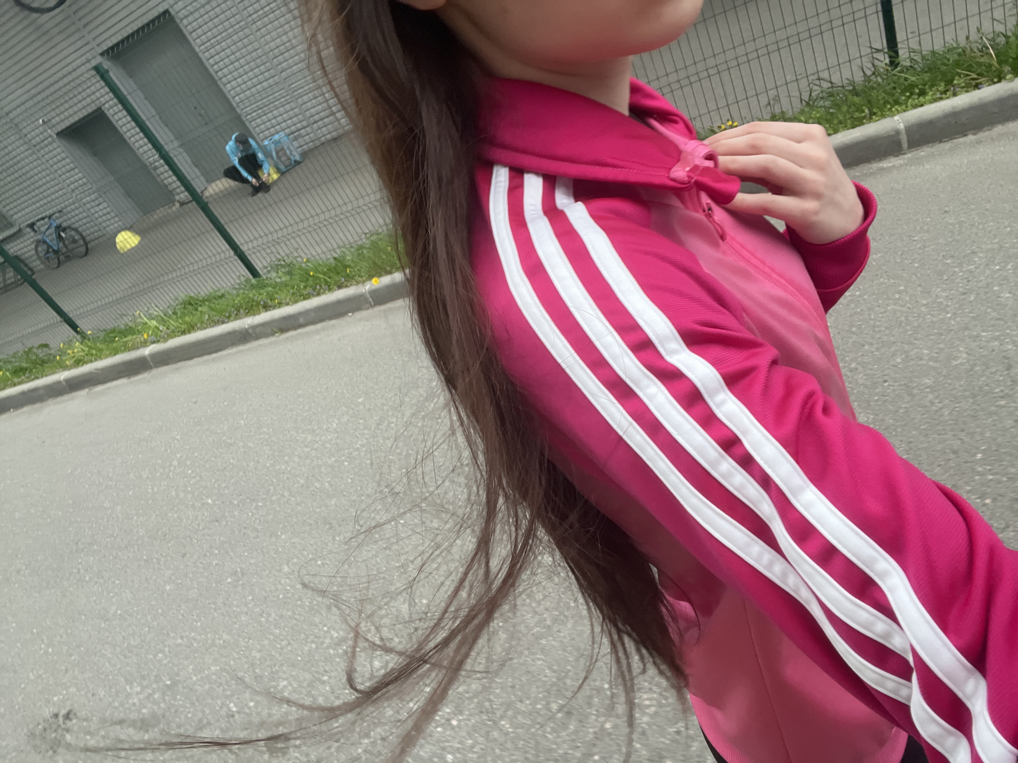 A person wearing a pink tracksuit stands on a gray asphalt surface. The tracksuit has three white stripes down the sleeve. In the background, there is a green fence, a building with gray doors, a bicycle, and a person tying their shoes. The location is potentially in Saint Petersburg, Russia, but this is not definitively verifiable from the image alone.