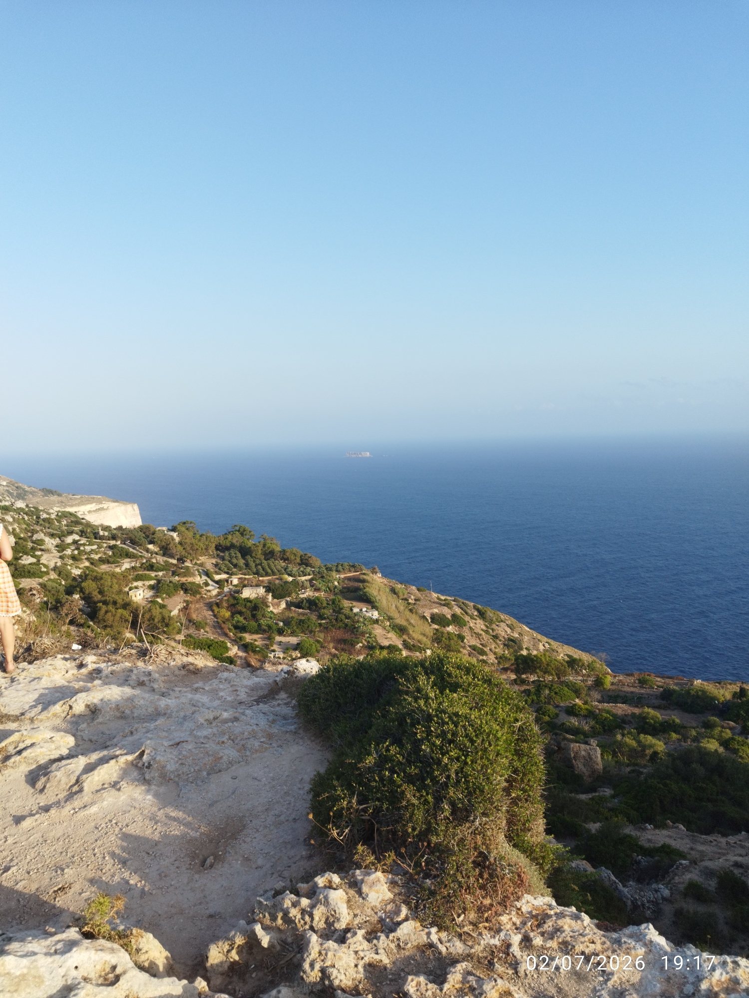 The image captures a scenic coastal view from a high vantage point, likely Dingli, Malta, given the context. The vast expanse of the deep blue Mediterranean Sea dominates the right half of the frame, meeting a clear, pale blue sky at the horizon. A lone, distant ship is visible on the water, hinting at maritime activity.
The foreground is a rocky, uneven cliffside dotted with sparse vegetation, including a prominent bushy green shrub. A portion of a person's legs and feet, clad in a patterned skirt, is visible on the far left, suggesting they are standing near the edge of the cliff, perhaps admiring the view. The landscape slopes down towards the sea, revealing a rugged terrain with more scattered greenery and what appear to be remnants of small structures or ruins, suggesting a historical or abandoned settlement on the cliff face.
The lighting indicates it is late afternoon or early evening, with the sun casting soft shadows and highlighting the textures of the rocks. The overall mood is serene and peaceful, emphasizing the natural beauty of the coastline. A timestamp is visible in the bottom right corner, reading "02/07/2026 19:17".