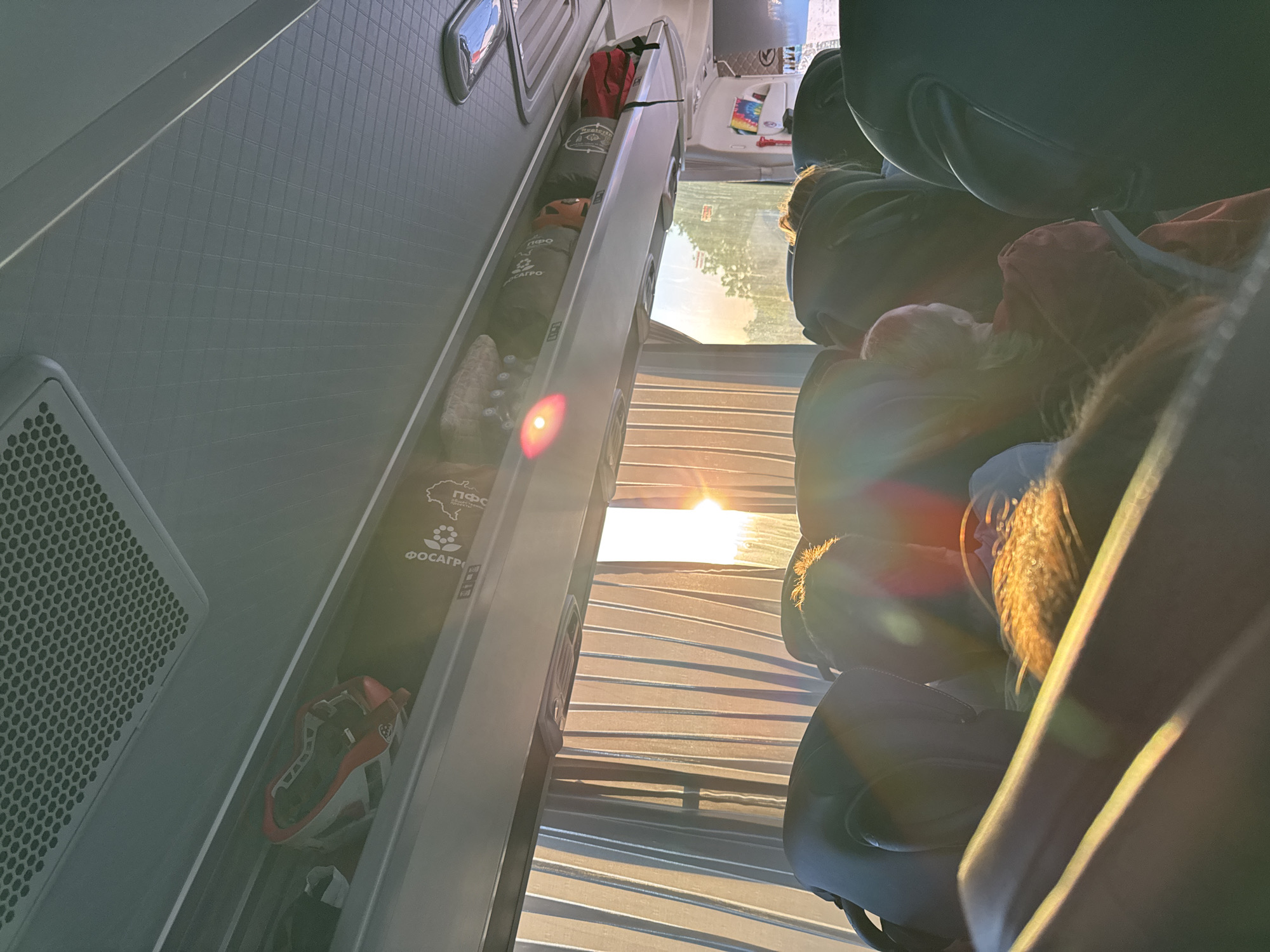 The image depicts the interior of a vehicle, possibly a camper van or RV. The view is from the back, angled towards the front. On the left is a storage compartment containing various items including a helmet, bags, and other travel gear. In the foreground, seats are visible. Through the vehicle's open blinds, a bright light source, likely the sun, shines through. Based on the presence of an item labelled "ФОСАГРО", which is a Russian agricultural company, the location could be in Russia, but not explicitly the city of Khvalynsk. The person is sitting. The environment appears to be outdoors, with the sun shining brightly.