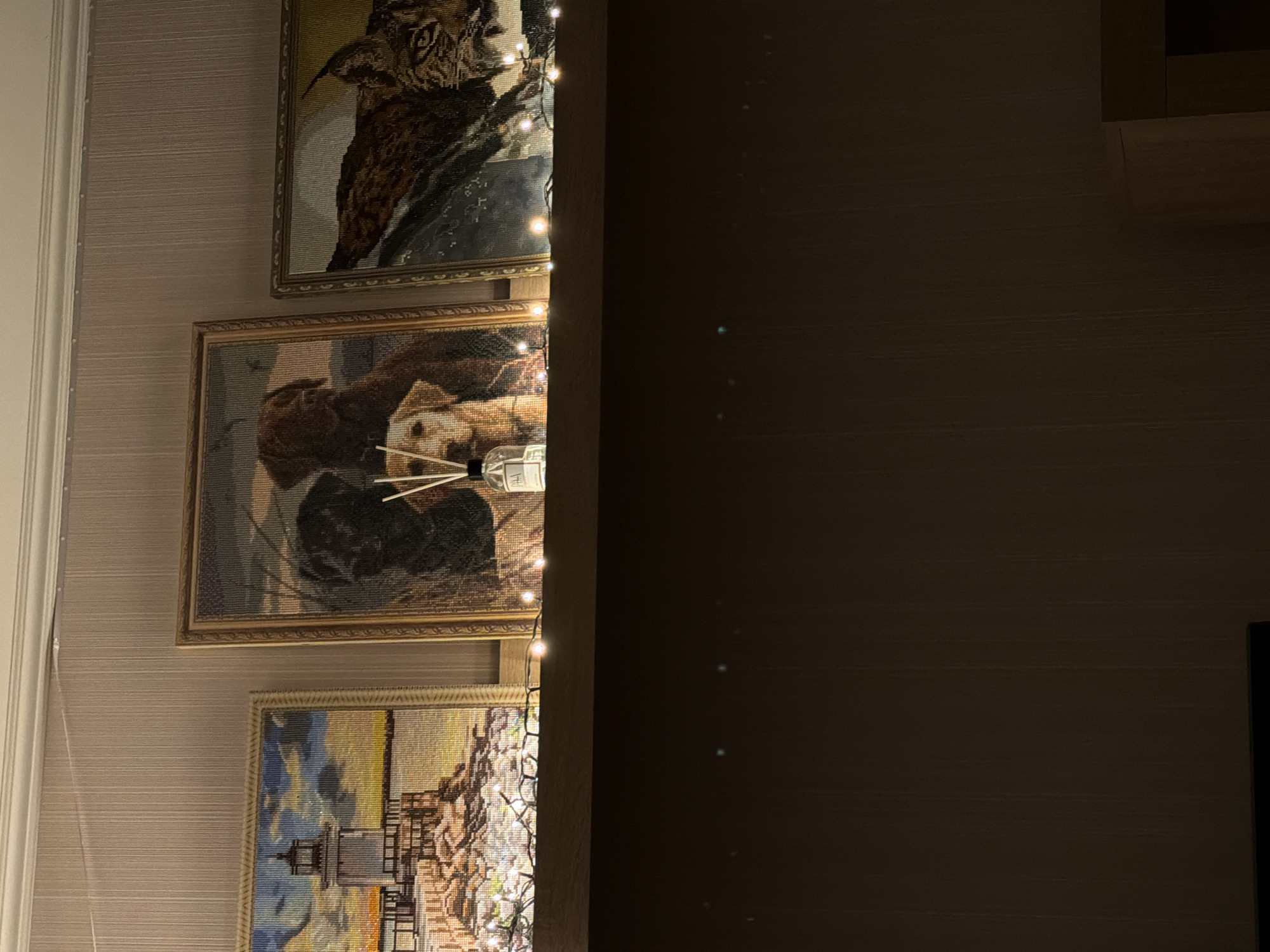 The image shows a shelf mounted on a wall with a textured wallpaper. On the shelf are three framed cross-stitch pictures. The leftmost picture depicts a lighthouse on a rocky shore with a body of water and a sky with clouds. The center picture is a portrait of three dogs: two black dogs and a golden retriever. In front of the center picture is a diffuser bottle with reeds. The rightmost picture features a lynx. A string of small, warm-toned fairy lights runs along the front edge of the shelf. The background wall appears to be a neutral color with vertical texture. The lighting in the scene is dim, with the fairy lights providing a soft glow. The overall setting suggests a home interior, likely a living room or hallway. There are no visible people or signs indicating a specific location in Cheltuitori, Moldova.