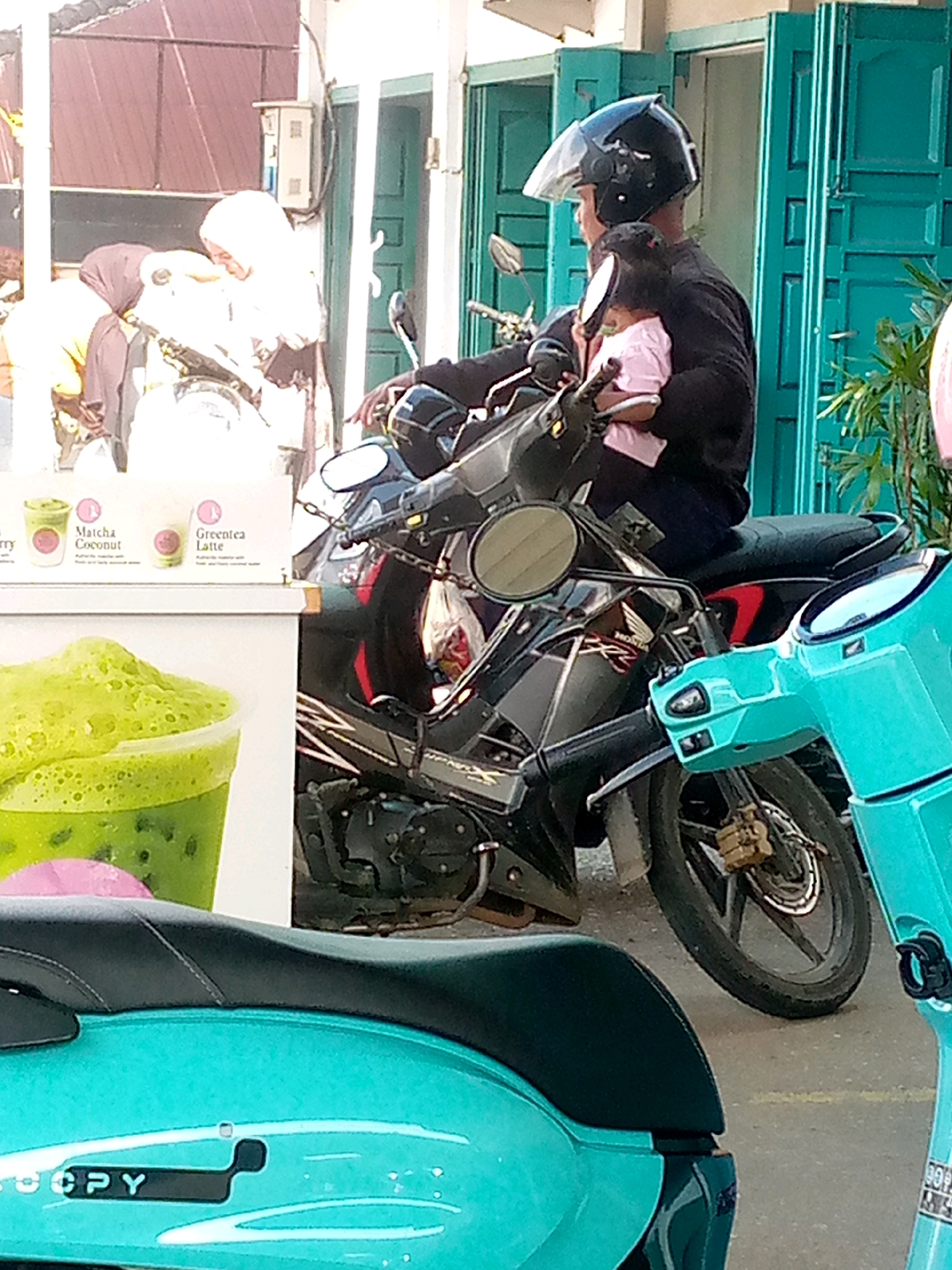 In Blangpeuria, Indonesia, a man wearing a black helmet and dark clothing is riding a black motorcycle. He is holding a young child, dressed in a pink outfit, on his lap. The motorcycle has chrome accents and a black seat. To the left of the motorcycle is a display board with images of drinks, including one labeled "Matcha Coconut" and another "Greentea Latte." In the foreground, a teal-colored scooter is partially visible, with the word "COPY" printed on its side. The background features turquoise doors and a building with a corrugated metal roof. Several other people are visible in the background, near other motorcycles and vehicles.