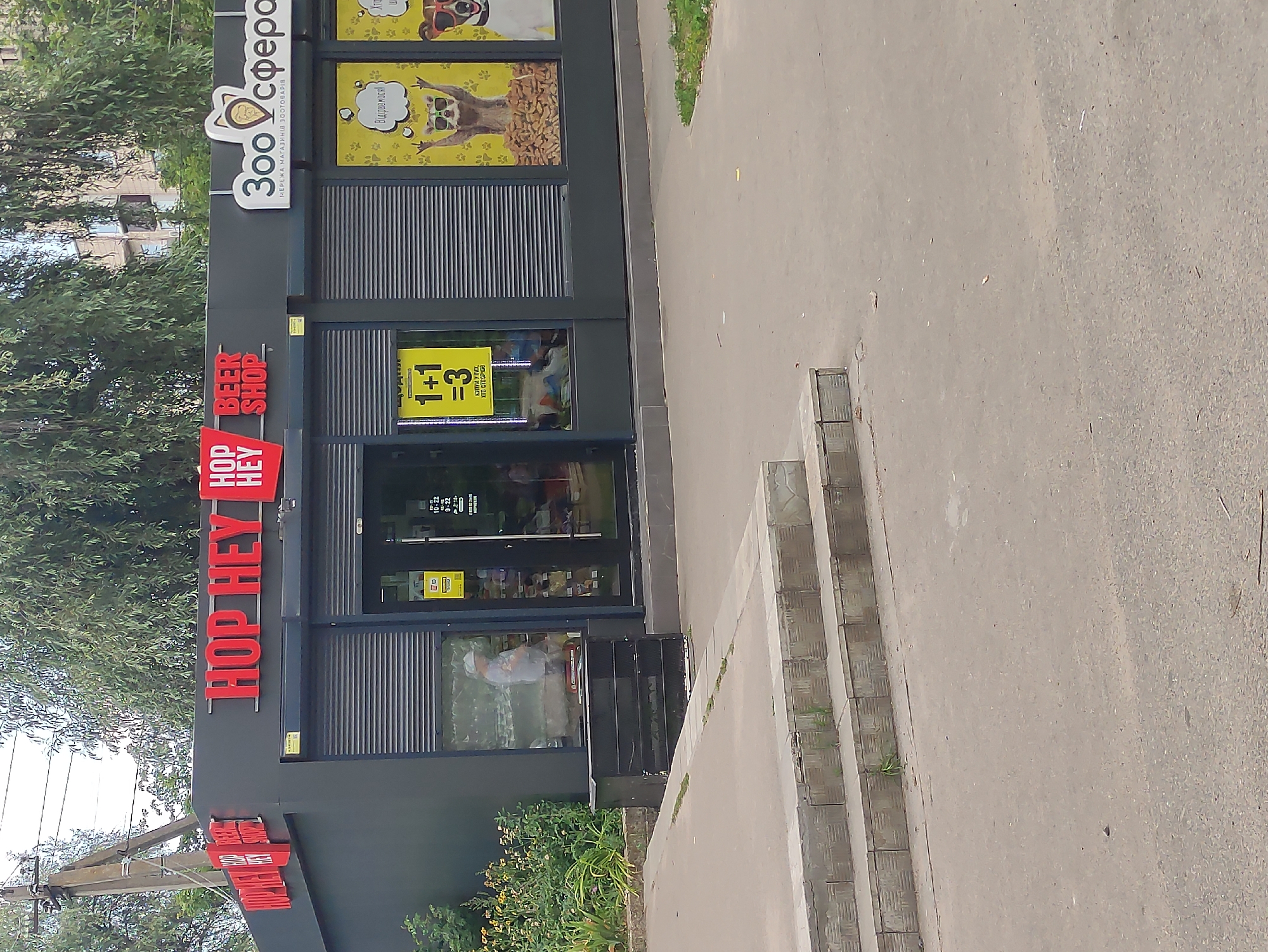 The image shows the exterior of two adjacent businesses in Boryspil, Ukraine. The business on the left is labeled "HOP HEY BEER SHOP" in large red lettering. The building is dark gray with rolling metal shutters covering the storefront. A yellow sign with "1+1 =3" is visible in the window. To the right, a pet supply store called "Зоосфера" (Zoosphere) is depicted. This store also has large red lettering and rolling shutters. The "Zoosphere" sign features a stylized cat logo. Yellow posters with cartoon animals are displayed in the windows. The storefronts are paved with concrete, and a set of concrete steps leads down to the sidewalk. Green trees and foliage surround the buildings, and a utility pole is visible to the left.