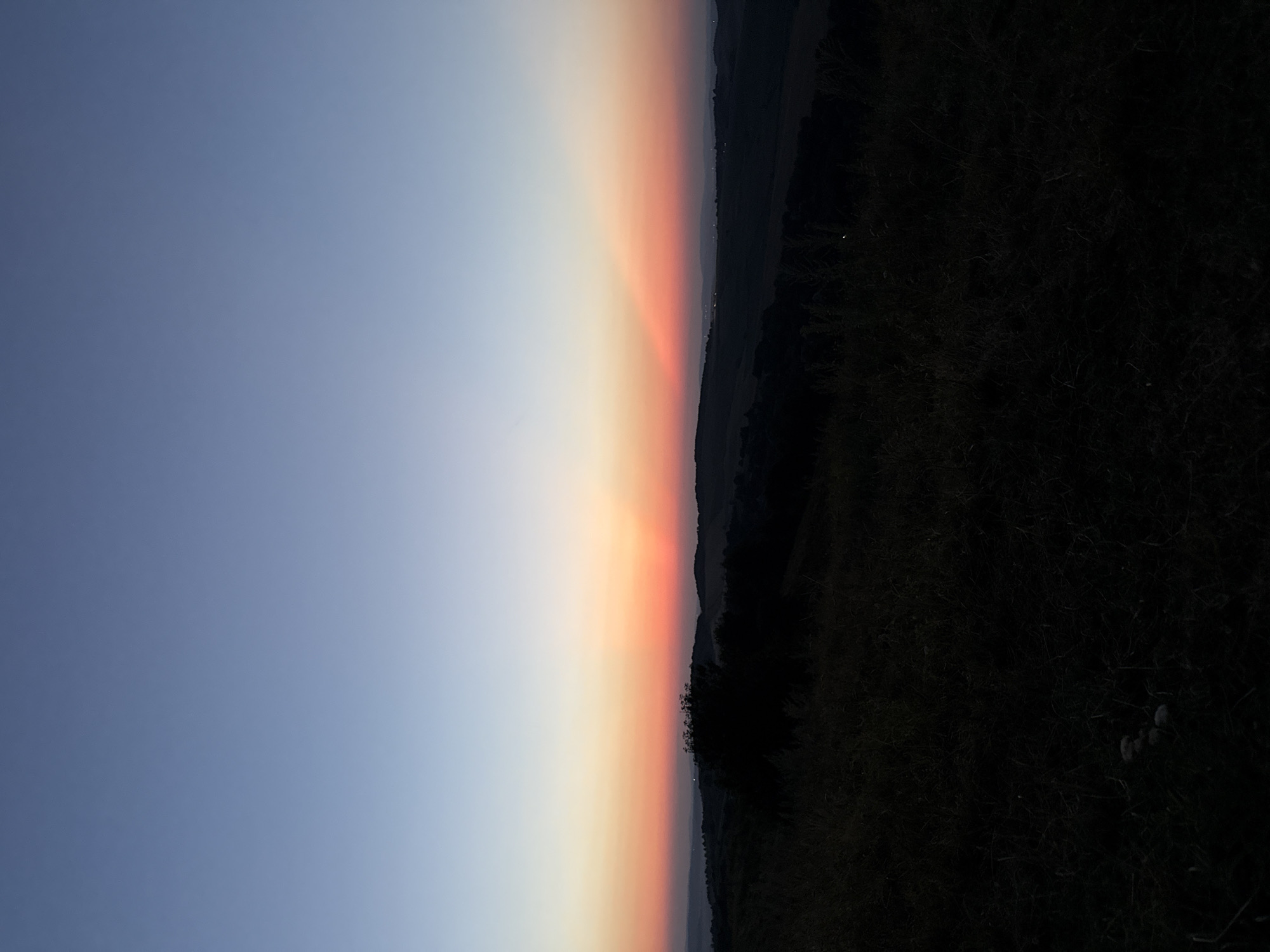 This is a photograph of a sunset or sunrise over a rolling landscape. The sky is a gradient of colors, transitioning from a pale blue at the top to a vibrant orange and pink near the horizon. Rays of light are emanating from a point on the horizon, creating a dramatic effect. The foreground is dark and silhouetted, showing the outlines of hills and vegetation, possibly a field of grass. A cluster of mushrooms can be seen in the lower left corner of the image. There are no people or animals visible in the scene. The overall impression is one of natural beauty and tranquility.