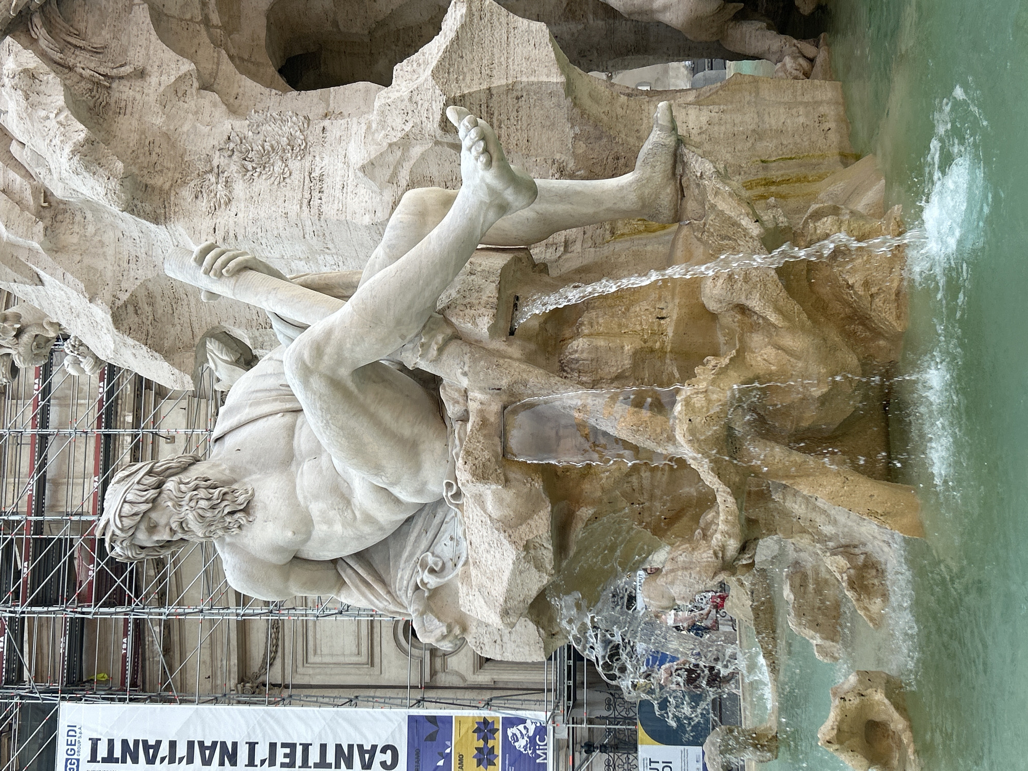 The image shows a close-up view of the Fountain of Four Rivers in Piazza Navona, Rome, Italy. The fountain features a large marble sculpture of a reclining male figure, likely representing the Nile River, with a beard and a laurel wreath on his head. He is seated on a rock formation from which water is flowing down into the pool below. His left leg is bent at the knee, and his right leg is extended. He is holding a cylindrical object in his right hand. In the background, scaffolding and a large banner with Italian text are visible, indicating ongoing construction or maintenance work. The water in the fountain pool is a clear turquoise color. Several people are visible in the background, some wading in the water, others standing near the edge of the fountain. The overall scene depicts a majestic architectural feature of Rome, partially obscured by scaffolding, with the refreshing element of flowing water.