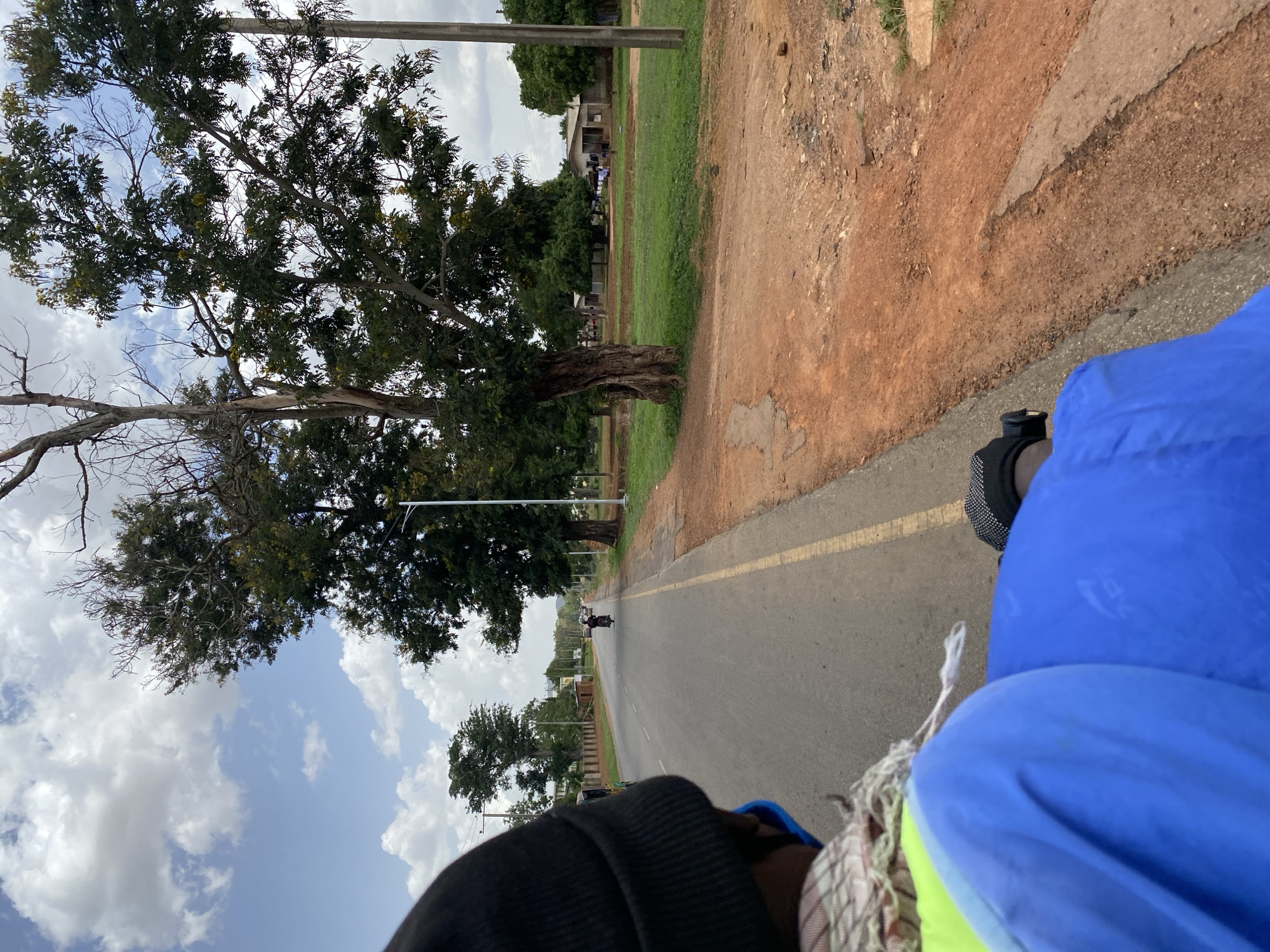 A low-angle, point-of-view shot shows an asphalt road stretching into the distance in Unknown, Nigeria. A yellow dividing line runs down the middle of the road. On the right, a dirt shoulder transitions into green grass. Trees line the left side of the road against a backdrop of blue sky with scattered clouds. In the distance, a motorcyclist is visible. In the immediate foreground, the blurred image of a person wearing a blue jacket and a black shoe is captured.