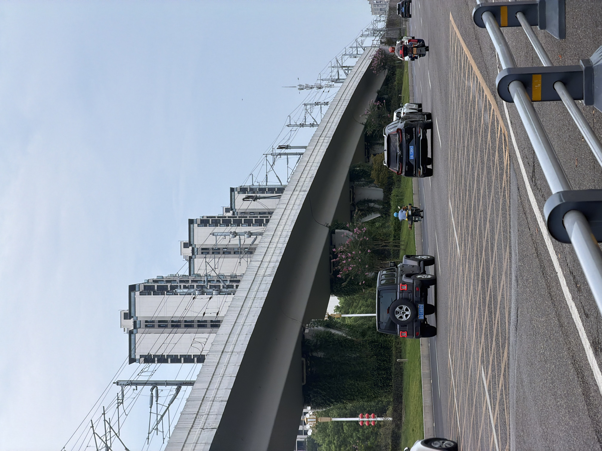 The image depicts a street scene in Jiangjin, China, under an elevated viaduct. The viaduct, a long concrete structure, carries overhead power lines supported by numerous poles. Several modern, multi-story apartment buildings are visible in the background. On the street below, a black Jeep Wrangler is in the foreground, facing away from the camera. To its right, a black SUV and a white car are also visible. Further down the road, two motorcycles are traveling in the same direction as the cars. One motorcycle has a rider wearing a red helmet, and the other has a rider in a blue helmet. The street is paved with asphalt and marked with yellow and white lines, indicating lanes and intersections. The scene is illuminated by overcast daylight.