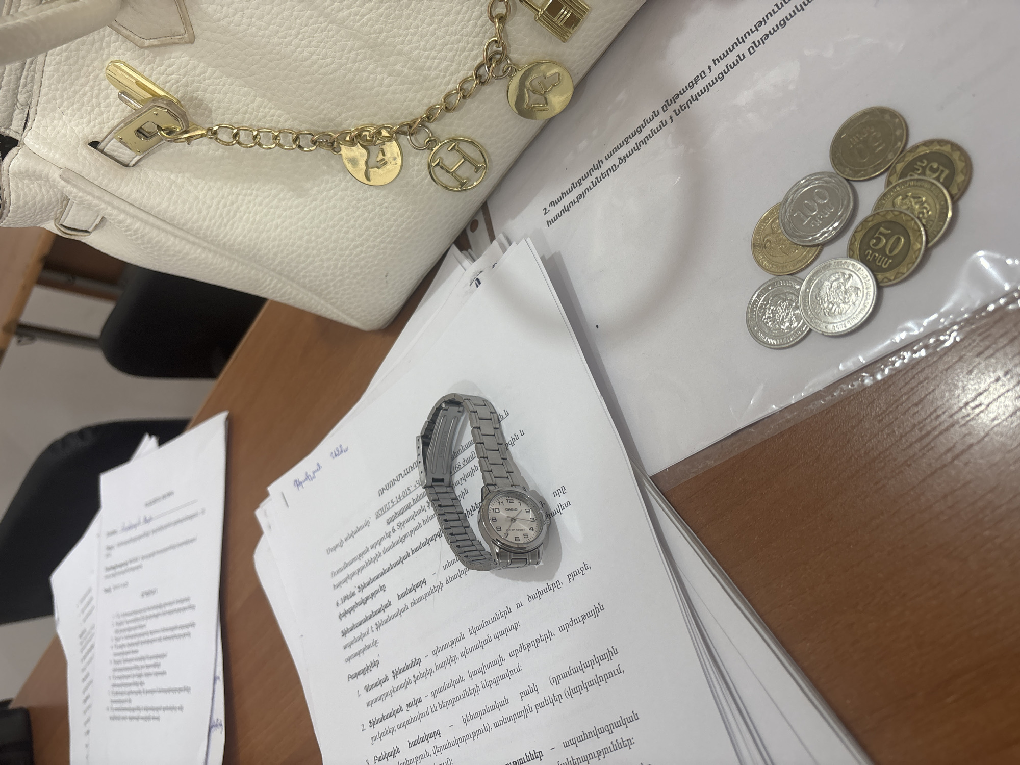 The image depicts a desk setting, likely in a university or office environment in Yerevan, Armenia. A white leather handbag with a gold chain adorned with charms is positioned at the top left. Stacked papers with Armenian text are in the center of the frame, upon which rests a silver Casio wristwatch. To the right, there is a small collection of Armenian coins of various denominations: 50, 100. The desk surface is a light brown color. A dark-colored office chair is partially visible in the background. The scene is well-lit.