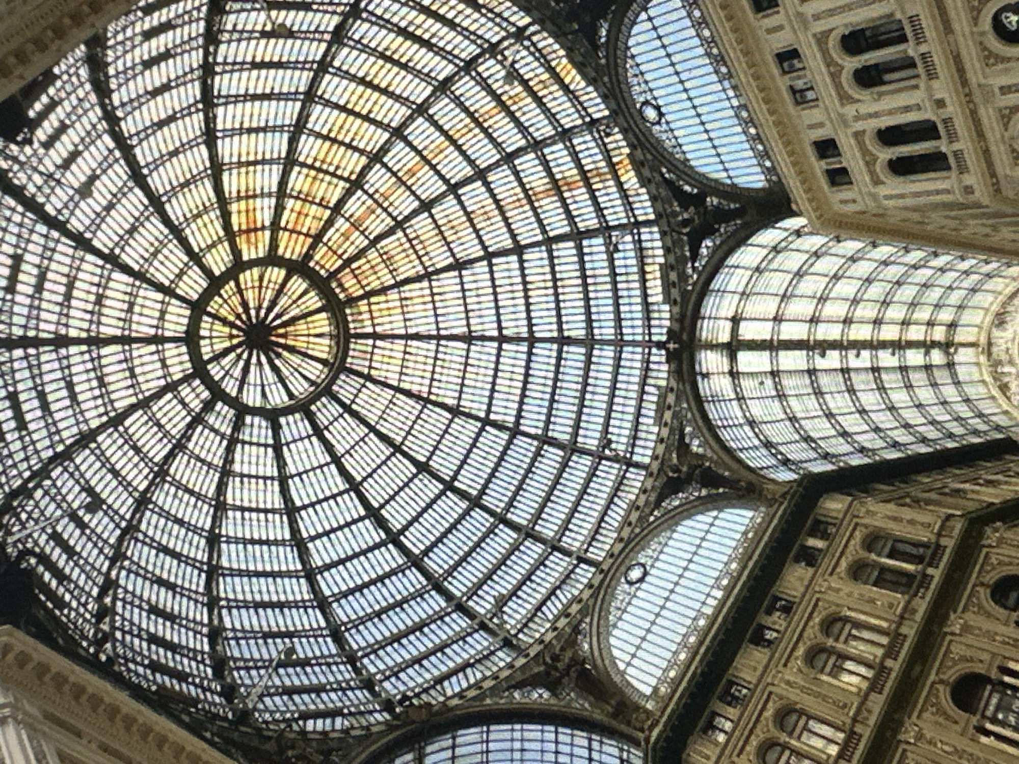 A wide-angle shot captures the interior of a grand arcade, characterized by an intricate, domed glass ceiling that arches overhead. The ceiling is constructed with a dark, skeletal framework of metal beams that divide the glass into a grid of rectangular panes, creating a geometric pattern against the bright sky. Sunlight filters through the glass, casting a warm, golden hue in some sections, while other areas are bathed in the cool blue of the sky.
Below the ceiling, ornate building facades rise up on either side of the arcade. These buildings are richly decorated with classical architectural elements, including arched windows, columns, and detailed carvings. The facades are painted in a light, creamy color, adding to the sense of opulence.
The perspective is from a low vantage point, emphasizing the vastness and height of the structure. The image is dominated by the interplay of light and shadow, the geometric patterns of the ceiling, and the detailed architecture of the surrounding buildings. There are no people visible in the frame.