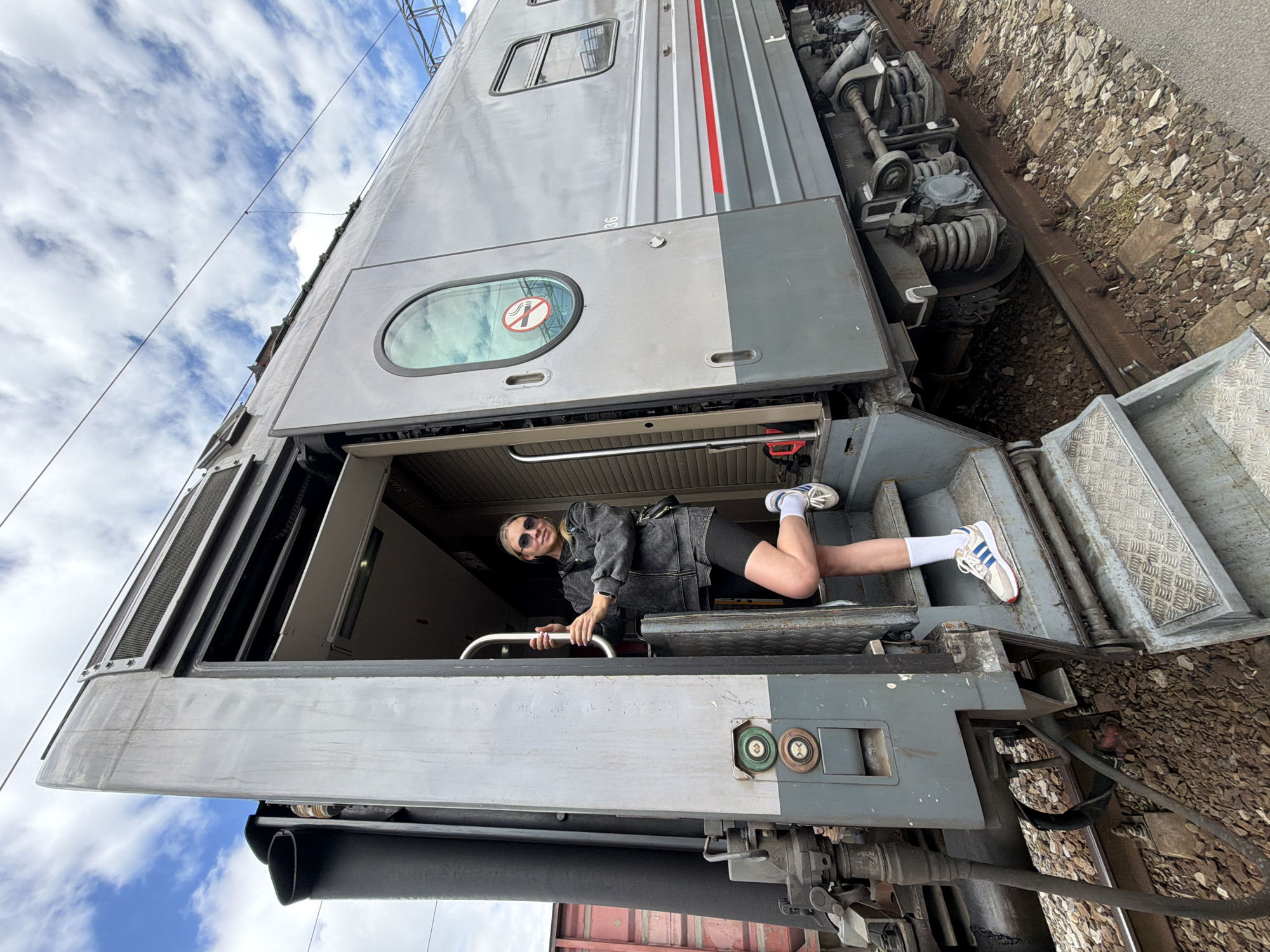 A woman is standing in the open doorway of a parked train carriage. She is wearing a dark denim jacket, black cycling shorts, white socks, and sneakers, and she has sunglasses on. She is holding onto a metal handrail inside the train. The train is silver with grey accents and red stripes. The train is parked on a track bed covered in gravel, and the sky with clouds is visible in the background. The image was taken in Rostov-on-Don, Russia.
