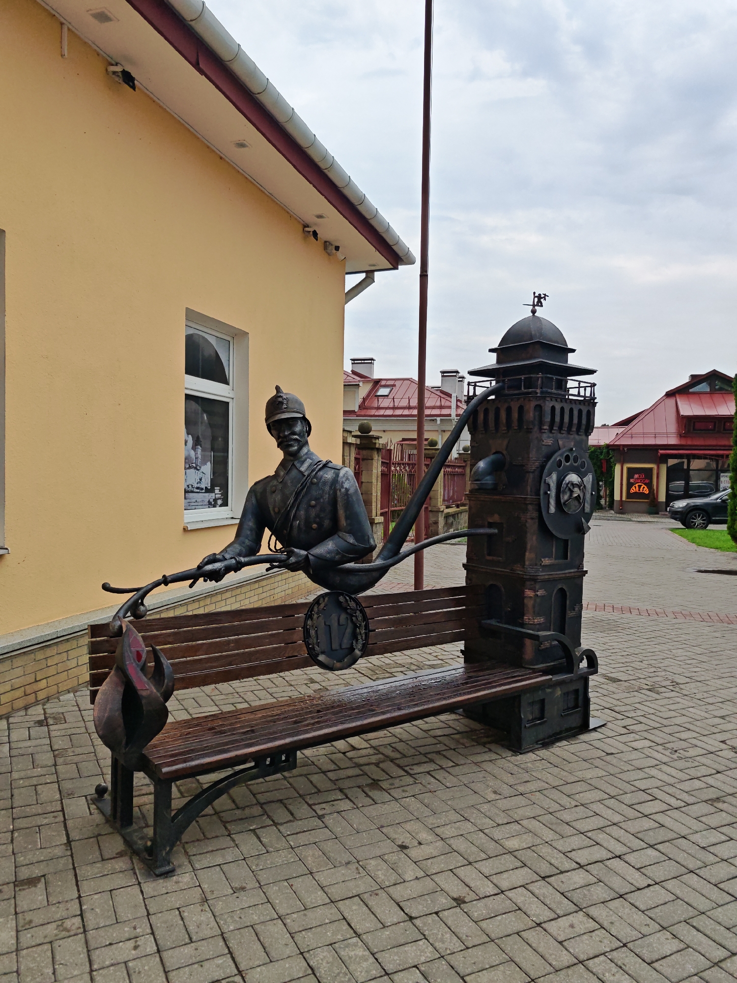 The image depicts a public art installation in the city of Hrodna, Belarus, likely near a fire station or a historical site related to firefighting. The sculpture features a bronze statue of a firefighter in uniform, complete with a helmet and mustache, leaning forward and holding what appears to be a hose. This sculpture is integrated with a park bench and a miniature fire tower. The fire tower is intricately designed, resembling a historical fire watchtower, complete with a clock face displaying the number "101," a common emergency number for fire services in some countries. The bench itself has decorative elements, including a flame motif at one end and a circular emblem with the number "12" on its backrest. The entire installation is situated on a paved walkway, with a yellow building in the background. The sky is overcast, suggesting a cloudy day. The context suggests this is a memorial or tribute to firefighters.