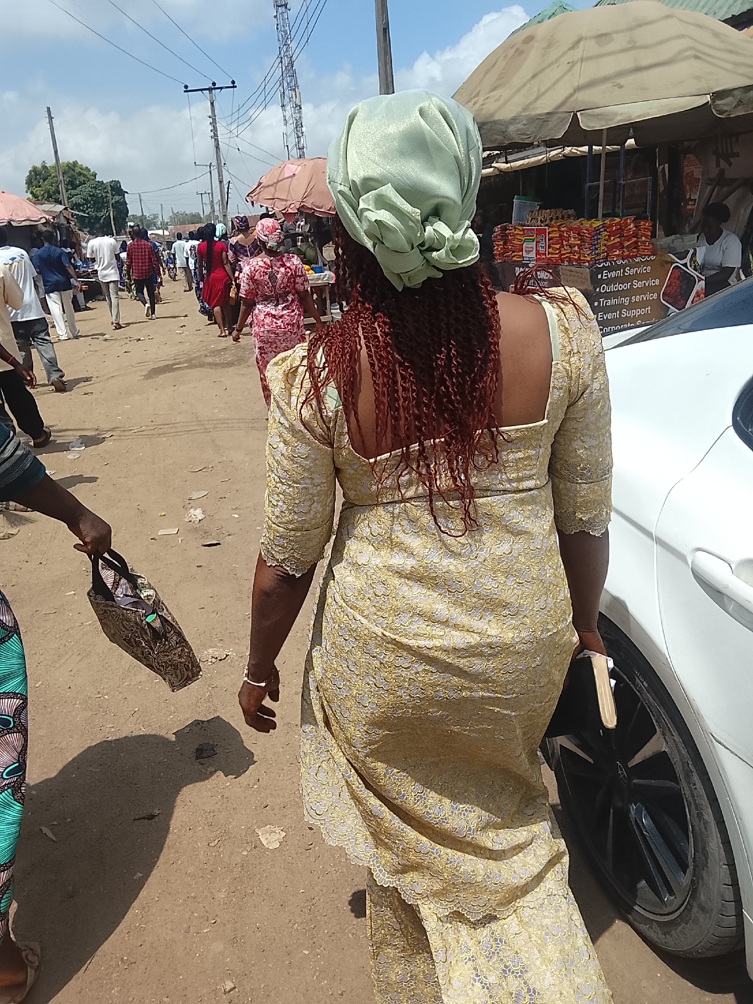 The image shows a street scene, likely a market, in Abuja, Nigeria. In the foreground, a woman with auburn twisted hair and a light green headwrap walks away from the camera. She is wearing a light gold, lace-patterned dress. To her left, another person is partially visible, holding a patterned handbag. To her right, a white car is parked.
The background shows a dirt road with other people walking in the same direction. Several market stalls are visible, indicated by umbrellas and structures with goods on display. Overhead, there are electrical wires and poles. The sky is bright with scattered clouds, indicating it is daytime. The scene is crowded and appears to be a busy commercial area.