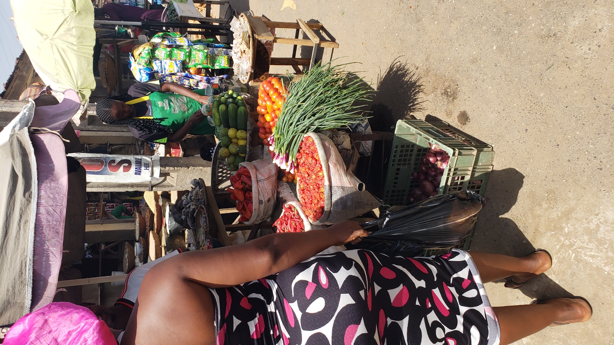 The image depicts a street market scene, likely in Narayi, Nigeria. A woman in a black and white patterned dress is seen from the knees down, holding a black plastic bag. Various vegetables, including tomatoes, onions, cucumbers, and lemons, are displayed for sale on wooden structures. Other vendors are visible in the background, along with goods such as snacks and other items under a light-colored canopy.