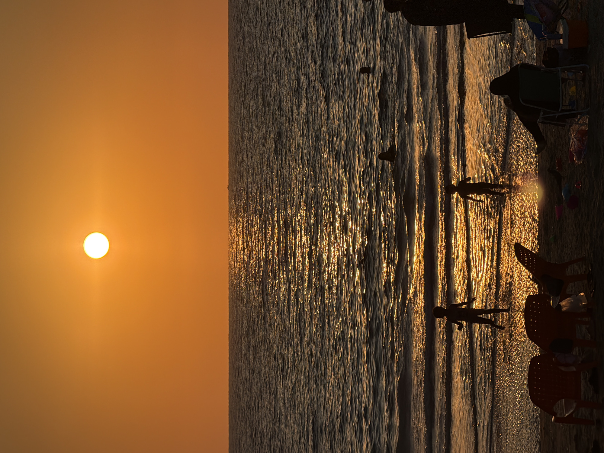 This media file depicts a serene sunset scene at a beach in Tel Aviv, Israel. The image is oriented vertically, with the sun prominently displayed as a bright orb on the left side, casting a warm orange glow across the sky and reflecting on the water. The sea is calm, with gentle waves lapping at the shore, creating shimmering patterns as the sunlight hits the surface.
Several individuals are present on the beach, silhouetted against the setting sun and the glistening water. Two children appear to be playing in the shallow water, their figures elongated by the low angle of the sun. Further back on the sand, several beach chairs, some orange plastic and one white foldable chair, are set up. A few people are seated in or near the chairs, also in silhouette, suggesting a relaxed atmosphere. The presence of beach toys scattered near the chairs hints at a family outing or a leisurely day at the beach.
The time of day is clearly sunset, indicated by the low position of the sun and the warm, golden hues of the sky. The weather appears to be clear and pleasant, conducive to beach activities. The scene evokes a sense of peace and tranquility, with the sounds of gentle waves and the soft light of the setting sun creating a calming ambiance. There is no visible text in the image.