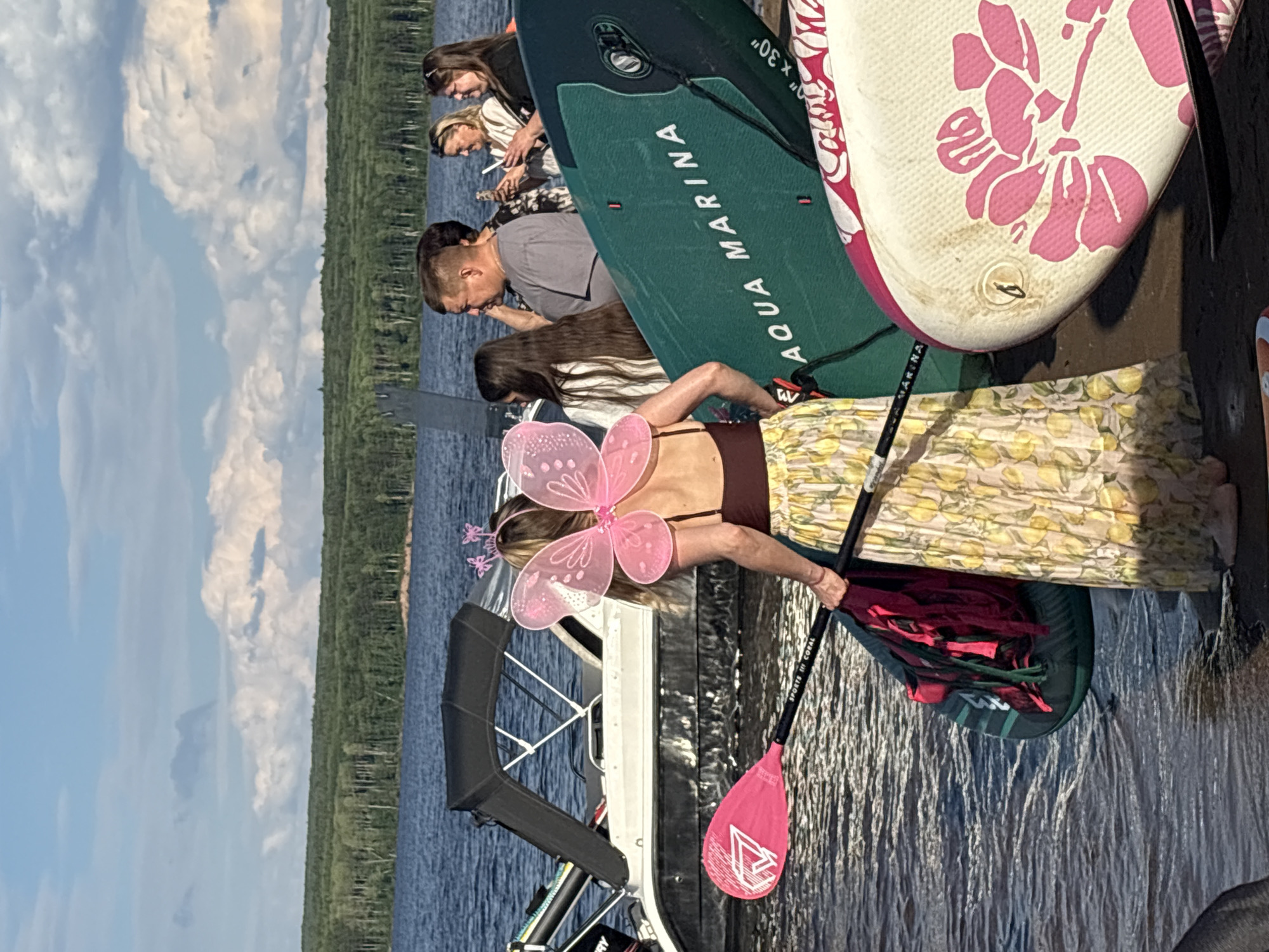 In Chaikovsky, Russia, a group of people are gathered near a body of water, likely the Kama River, on a sunny day. A woman in the foreground, dressed in a fairy costume with pink butterfly wings and a flower-patterned skirt, holds a pink paddle. She is standing next to a teal paddleboard with "AQUA MARINA" printed on it, and a white paddleboard with a floral design. A boat is docked behind them. The background features a dense forest and a blue sky with scattered clouds. The people appear to be preparing for or have just finished a water activity.