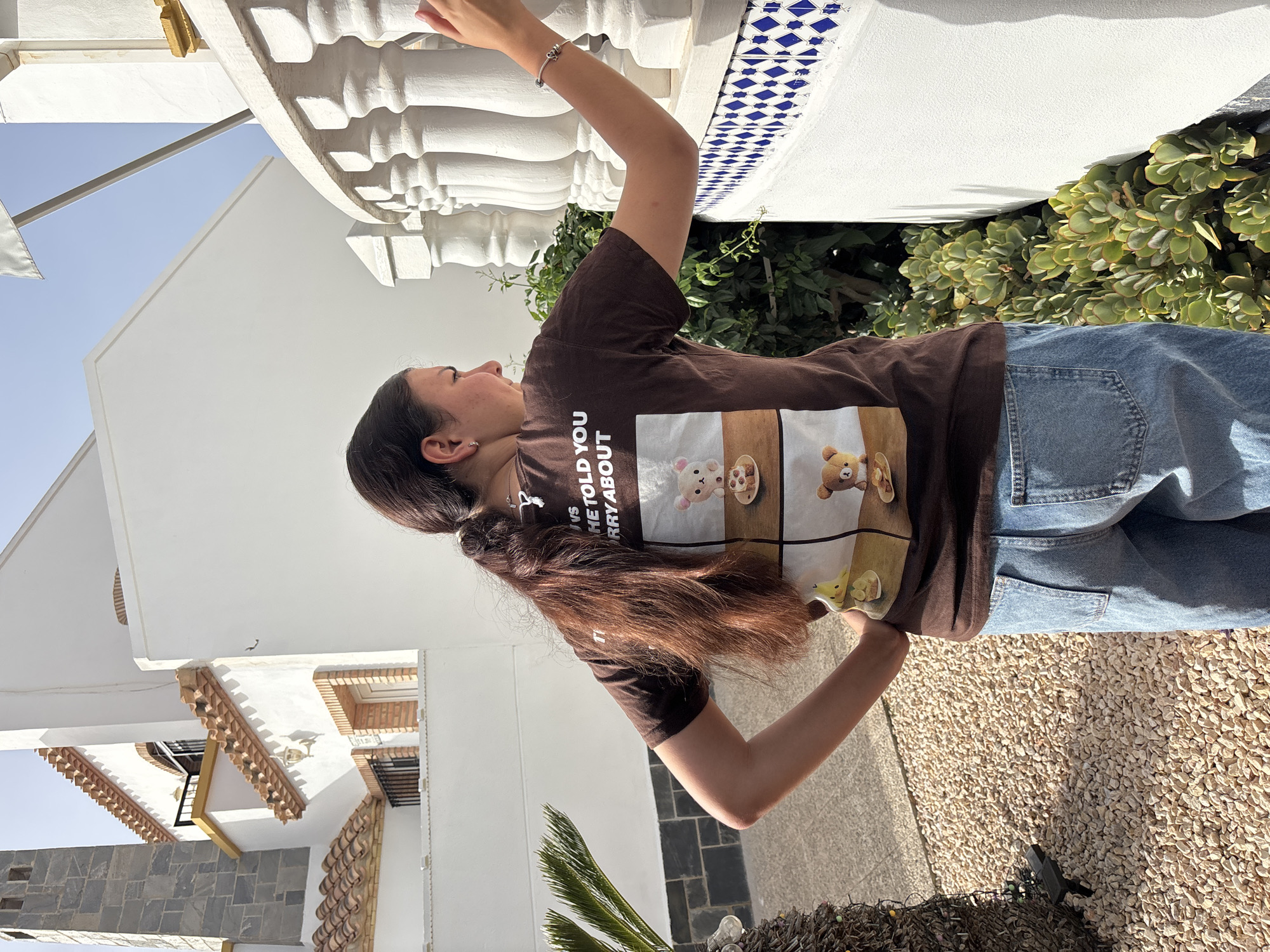 A young woman, viewed from the back and right side, stands outdoors. She has long, dark brown hair pulled into a ponytail. She is wearing a dark brown t-shirt with a graphic print of four panels, each featuring a cartoon bear character and food. She is also wearing light blue denim jeans. The woman is leaning her right arm on a white, ornate railing and is looking to her right. Her left hand is on her hip. The background consists of a white building with a tiled roof and orange trim, a grey stone chimney, and a small palm-like plant. The ground is covered in light-colored gravel. The overall setting appears to be a residential area, possibly a patio or balcony, in a sunny climate. The woman is standing near a building that is characteristic of architecture found in coastal areas of Spain, such as Orihuela, Spain, which has a Mediterranean climate with bright sunshine and whitewashed buildings.
